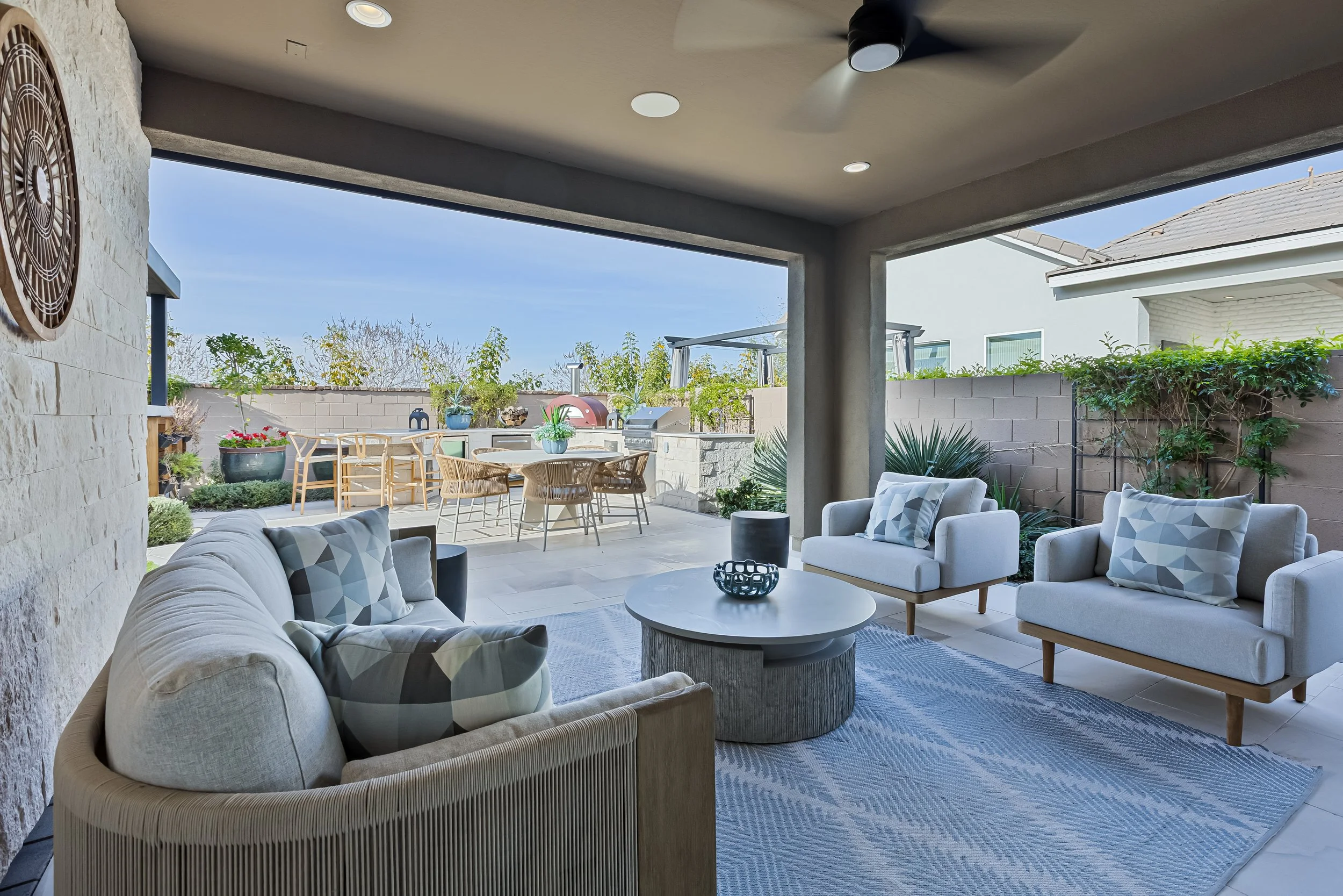 View of a modern outdoor patio with a seating area featuring light-colored sofas and chairs with geometric pillows, a round coffee table, a outdoor dining table with chairs, plants, and a built-in grill in the background, under a covered ceiling with