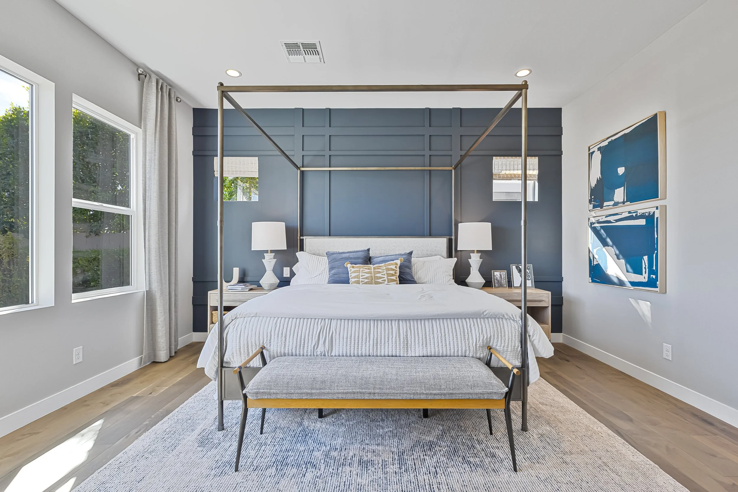 Modern bedroom with a bed under a four-poster frame, navy blue accent wall behind the bed, two nightstands with lamps, artwork on the walls, and large windows offering natural light.