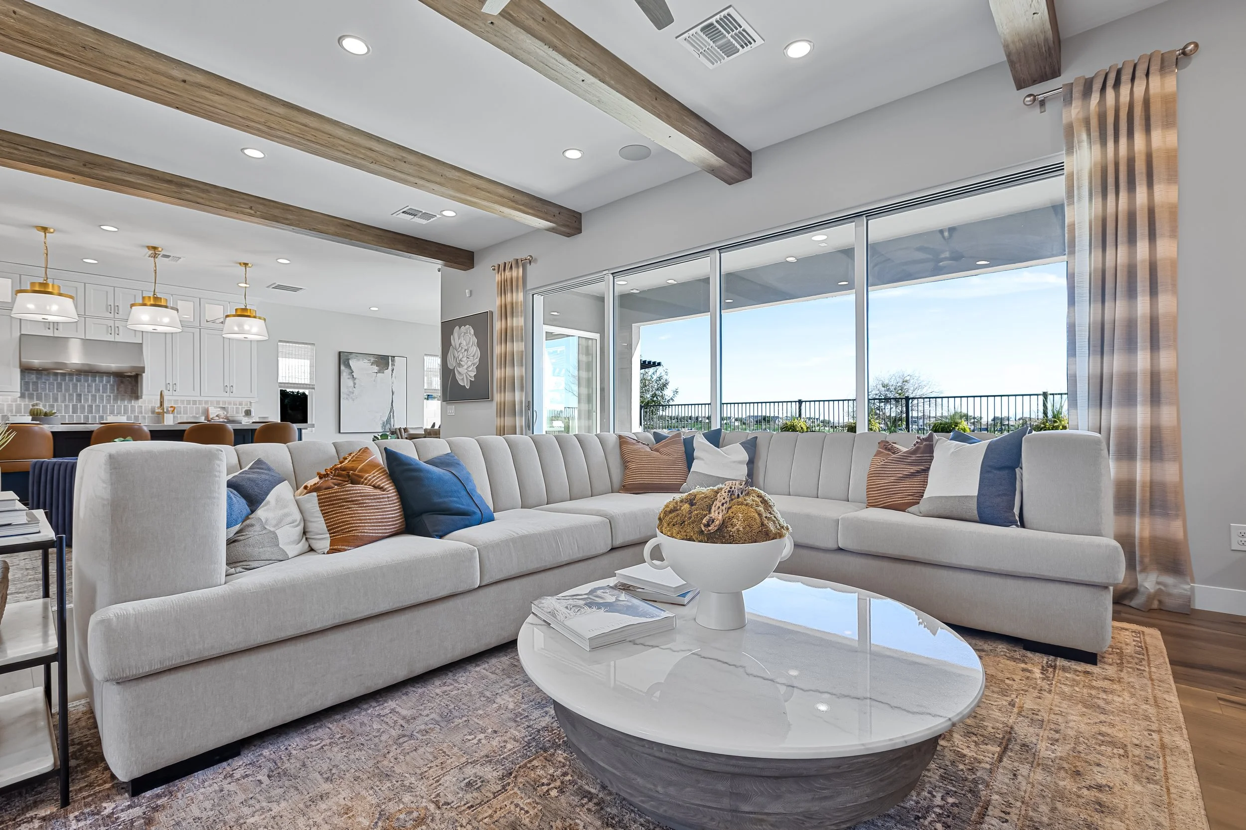 Living room with a large cream sectional sofa with striped and solid pillows, a white oval coffee table with books and a decorative bowl, large windows with beige curtains displaying an outdoor view, a kitchen area with white cabinets, hanging lights