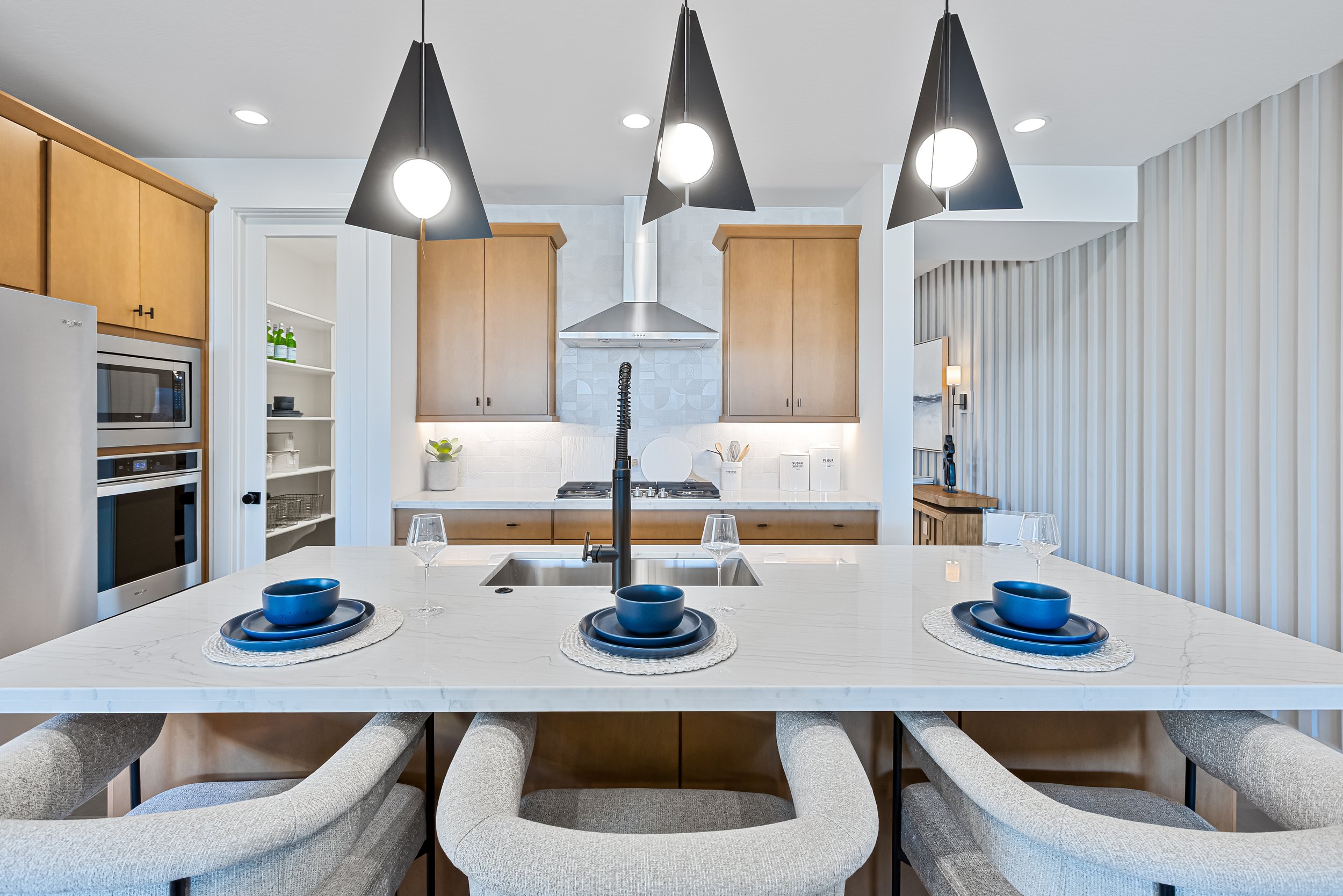 Modern kitchen with a white marble island, blue dishware, gray chairs, wooden cabinets, stainless steel appliances, and black pendant lights.