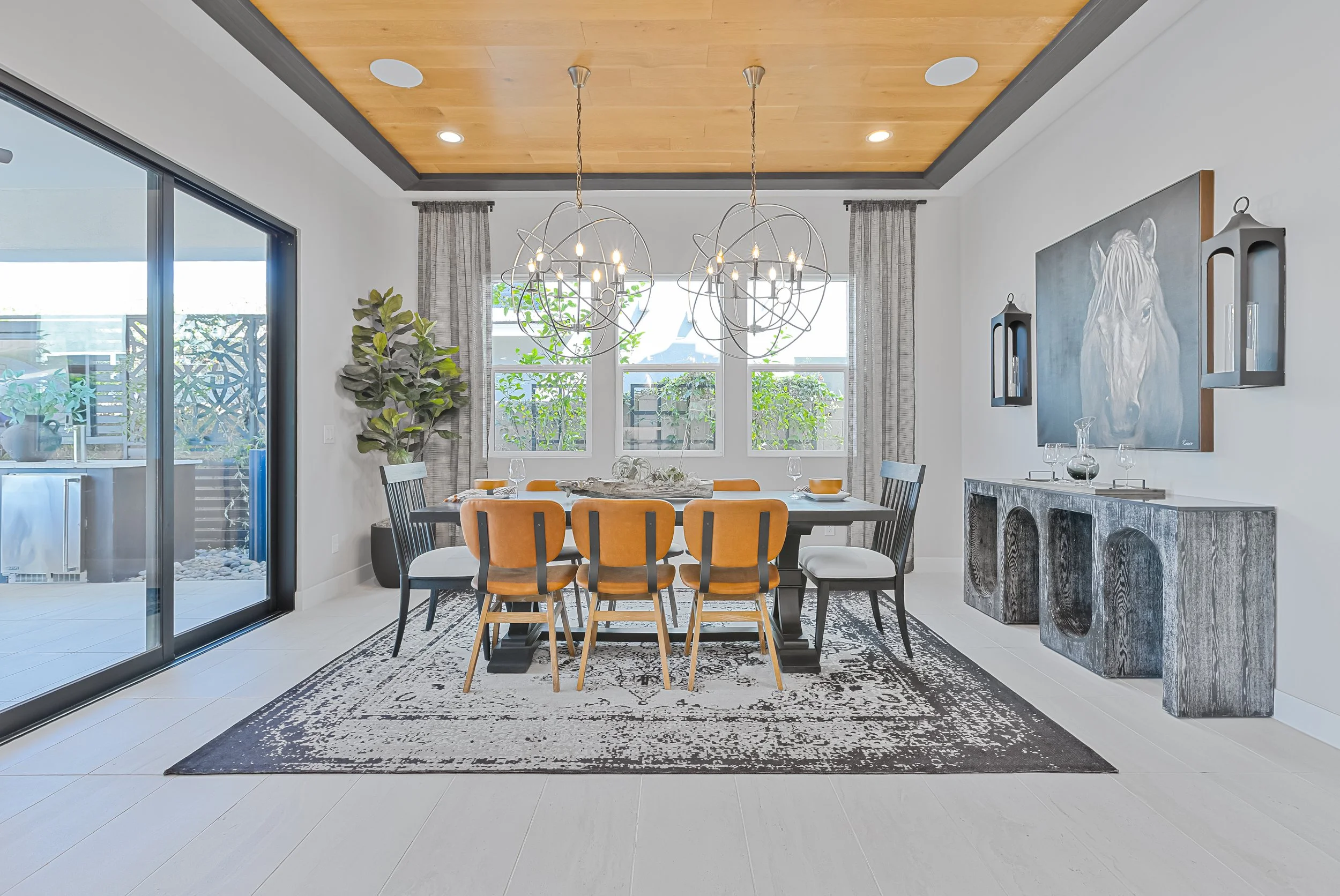 Modern dining room with a black and white rug, a large wooden table, and six chairs with yellow and black upholstery. Two large chandelier light fixtures hang above. There are three windows with curtains, a sliding glass door, a large painting of a w