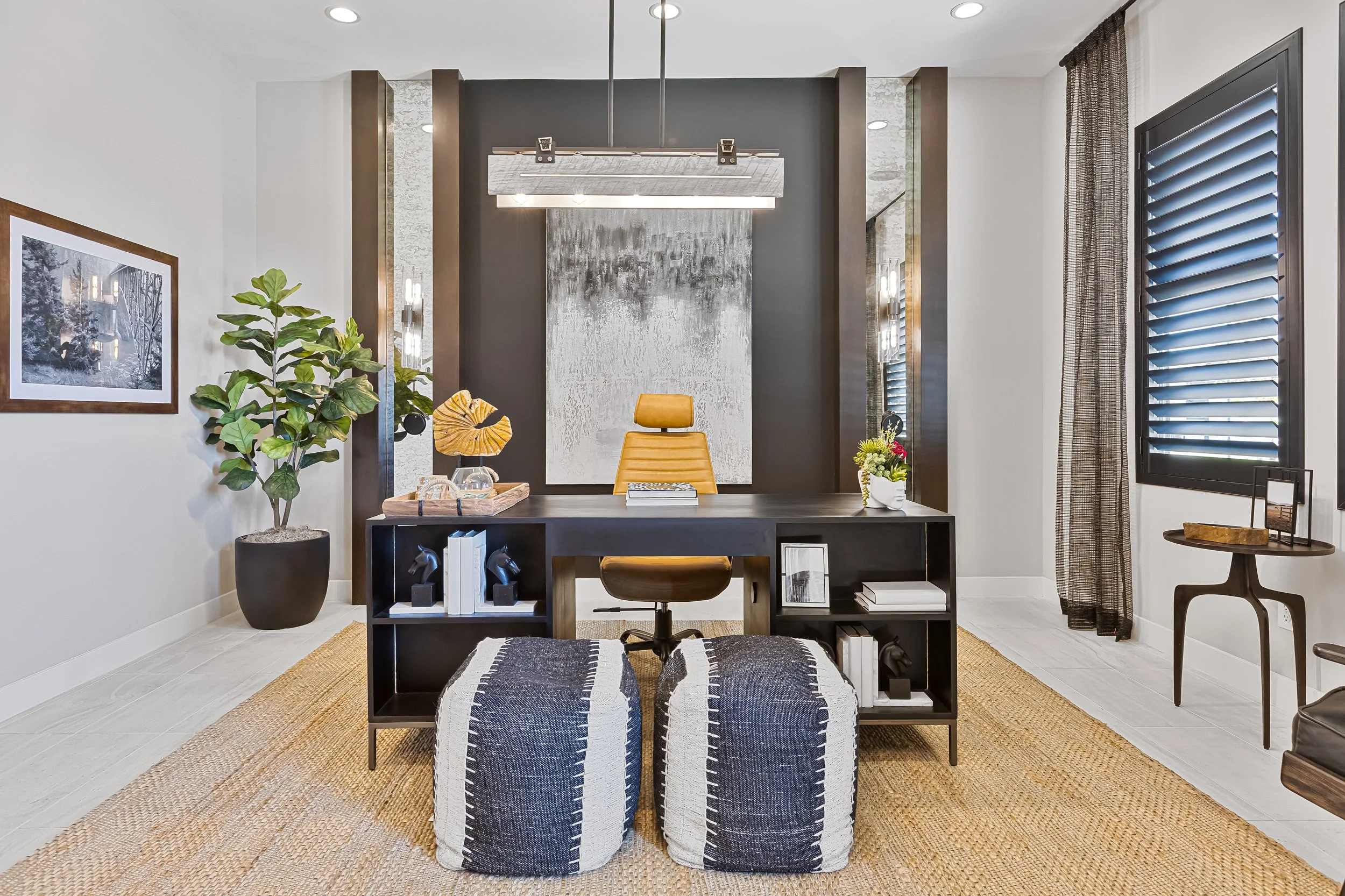 Modern office with a black desk, yellow office chair, and two black and white striped poufs, decorated with plants, artwork, and books, in a bright room with large windows and grey curtains.