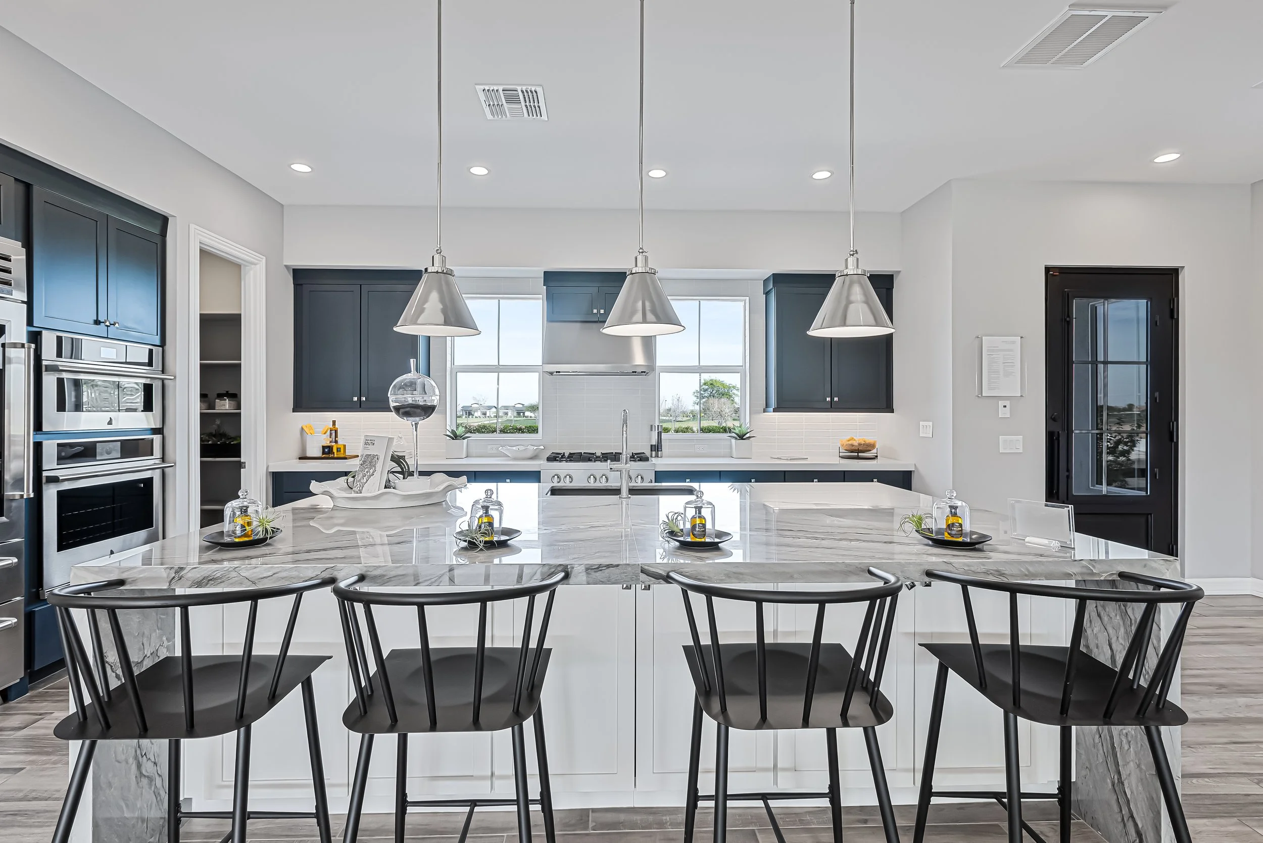 Modern kitchen with black cabinets, a marble island, three pendant lights, and stainless steel appliances, overlooking a bright window.