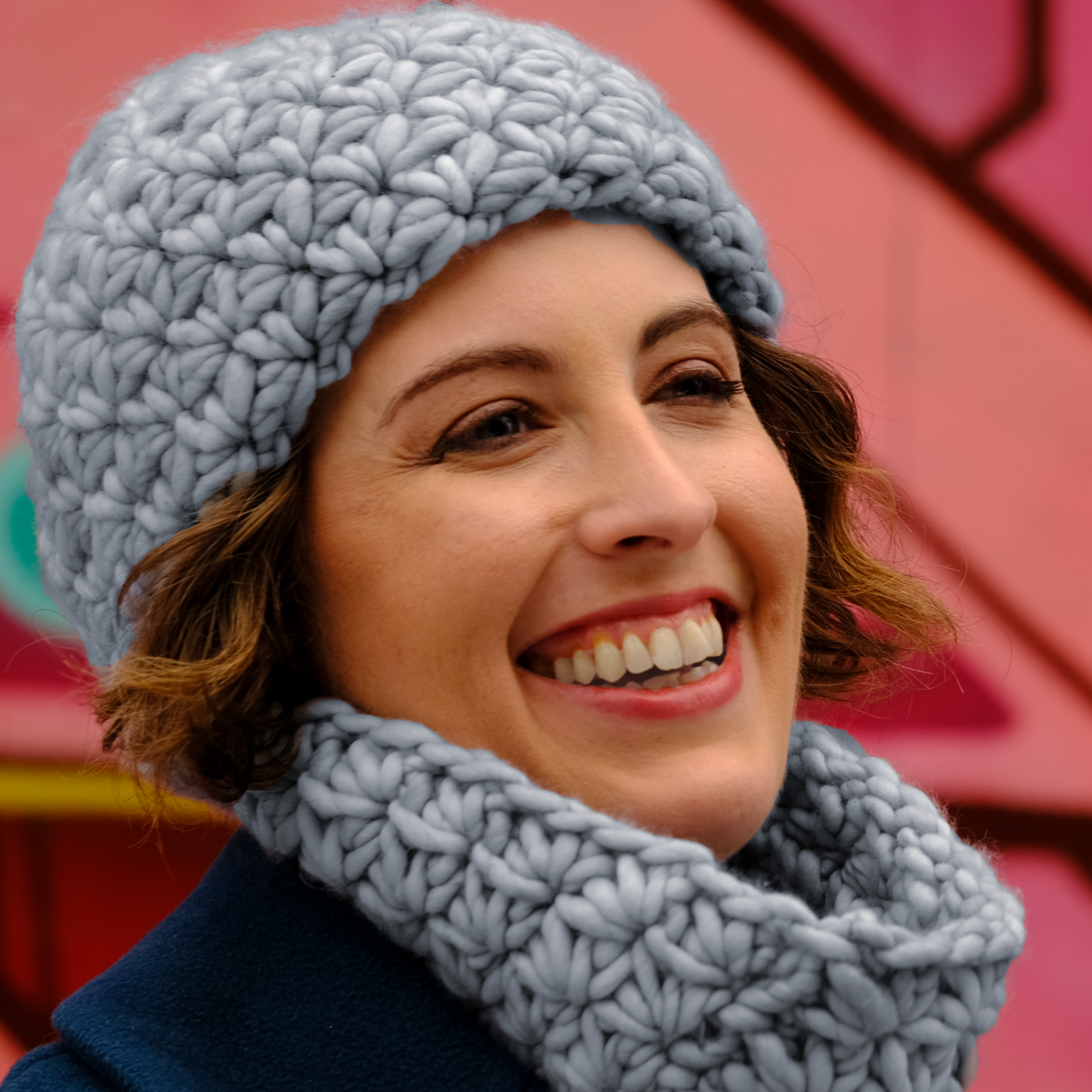 Close-up of a woman smiling, wearing a large, chunky light blue knit hat and a matching cowl, against a colorful mural