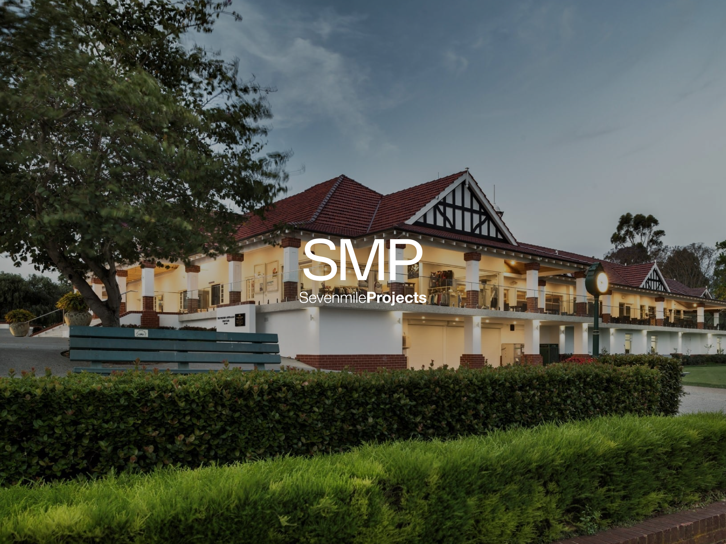 A large two-story residential building with a red-tiled roof, white exterior walls, and black decorative wood trim. It has a spacious balcony with outdoor furniture, surrounding green lawn, bushes, and trees, with a cloudy evening sky in the backgrou