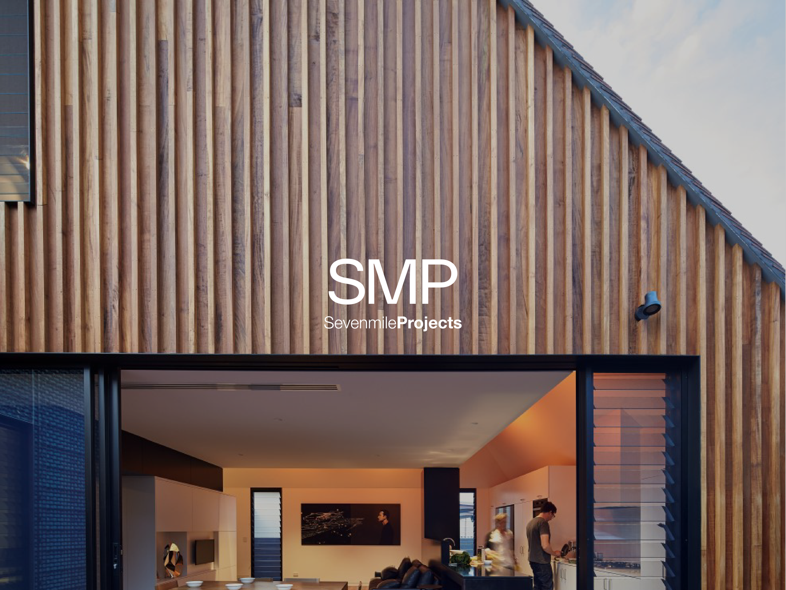 Photo of a modern house with wooden exterior panels and an open interior view, showing people inside in a kitchen and living room area.