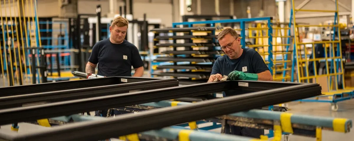 Two workers in a factory assembling window frames, with various shelving and carts in the background.