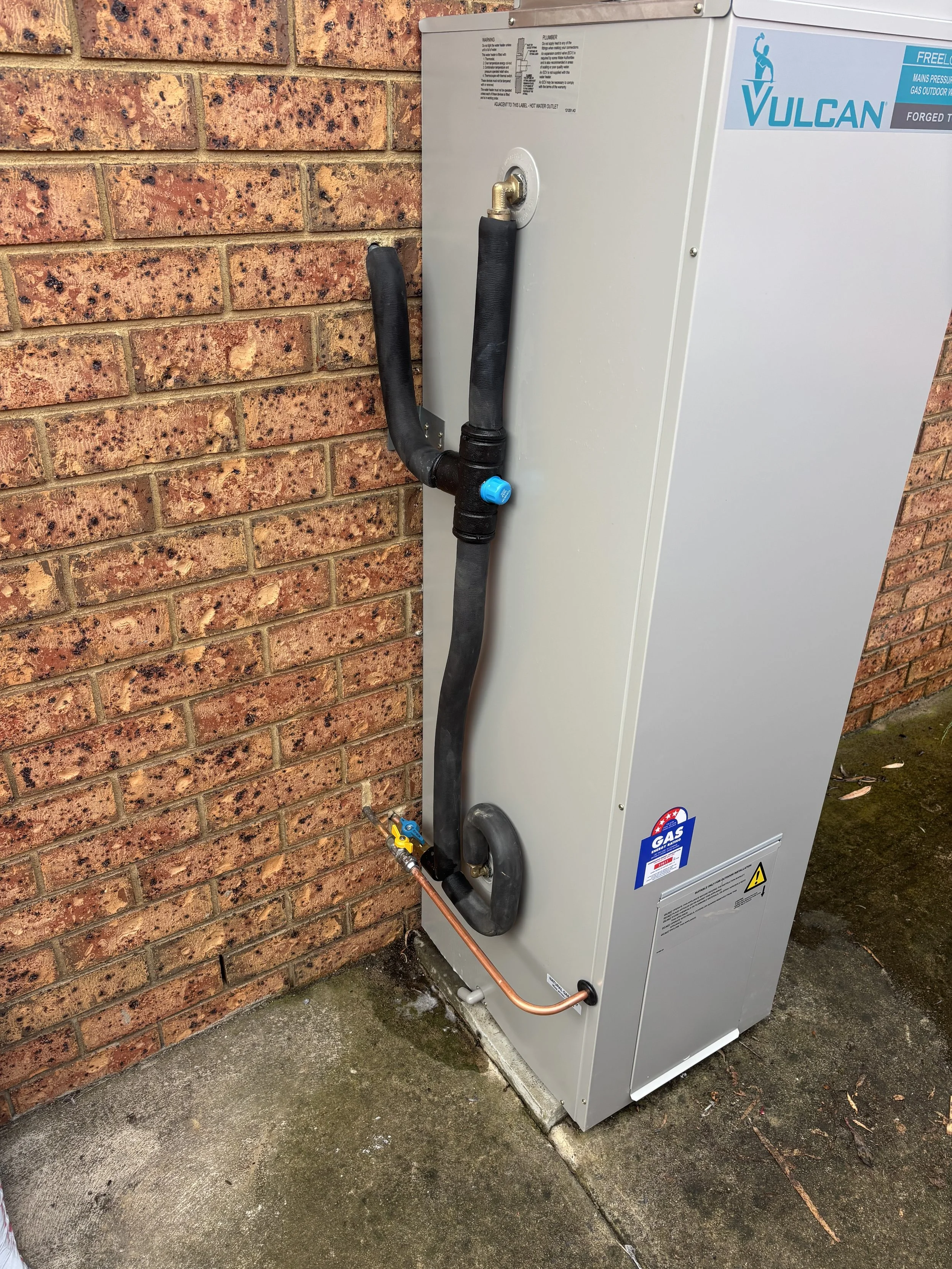 A Vulcan outdoor gas heating or water system unit mounted on a concrete slab outside a brick wall, with black pipes and copper tubing connected.