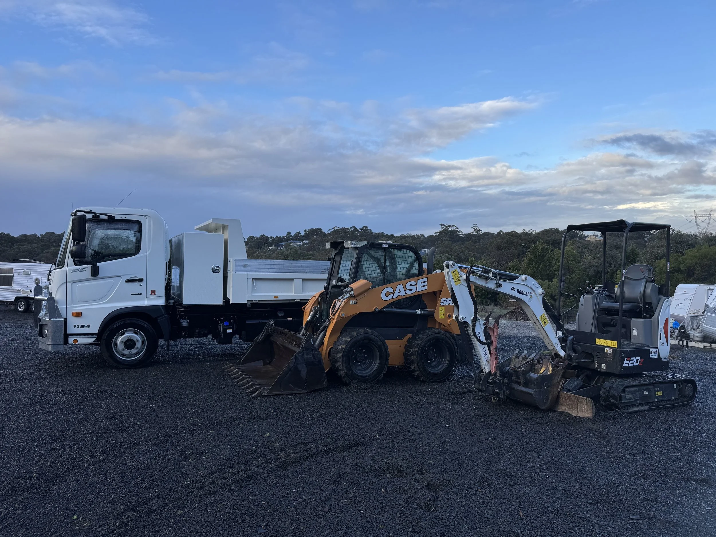 A white flatbed truck parked on a gravel lot, with a compact CASE skid-steer loader attached to it. The loader has a bucket attachment and is black and orange, with a transparent cab. The background shows a partly cloudy sky and some distant trees.