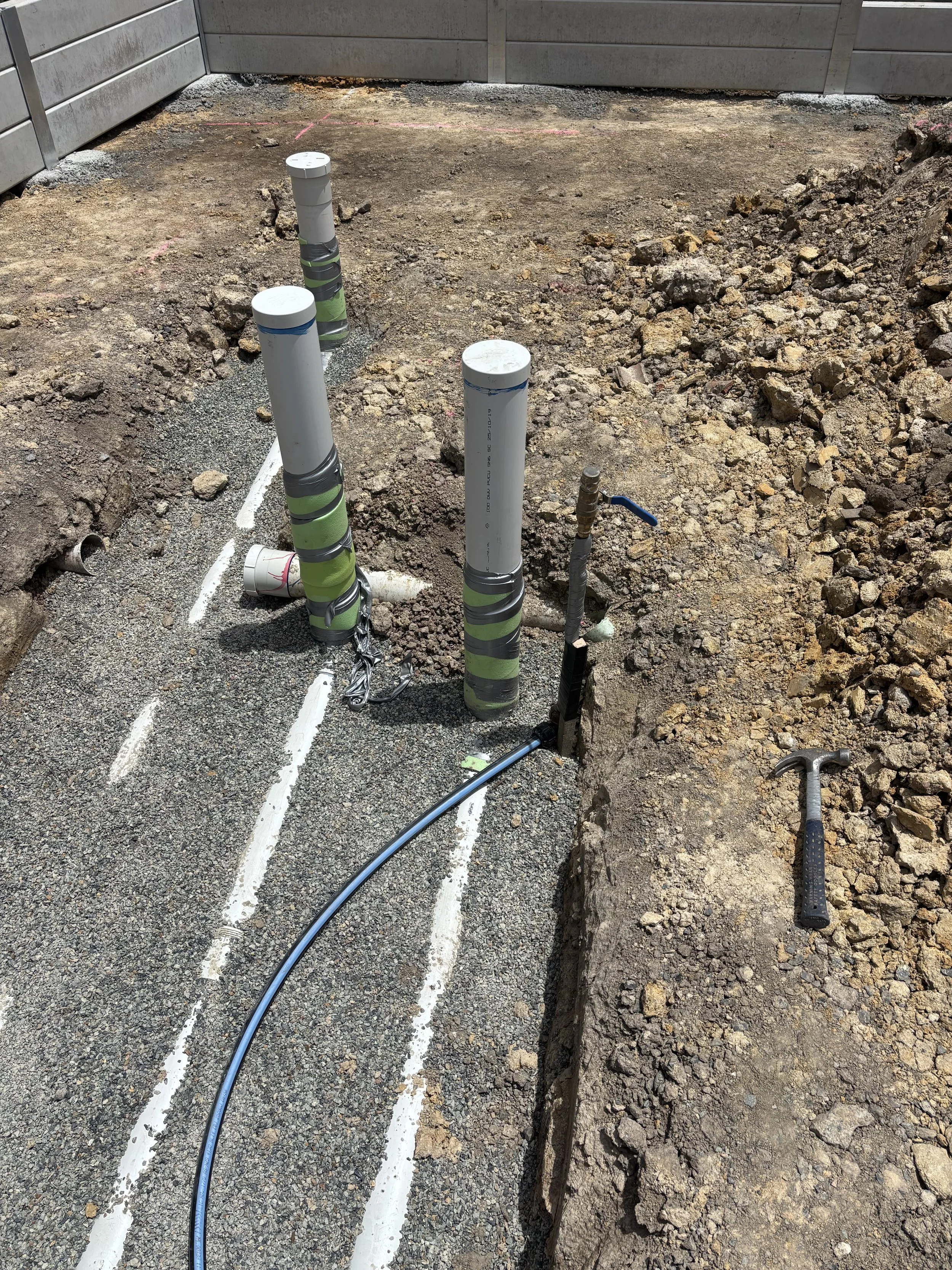 Outdoor construction site with underground plumbing pipes, some wrapped with tape, around a gravel base, and a hammer lying on the dirt.