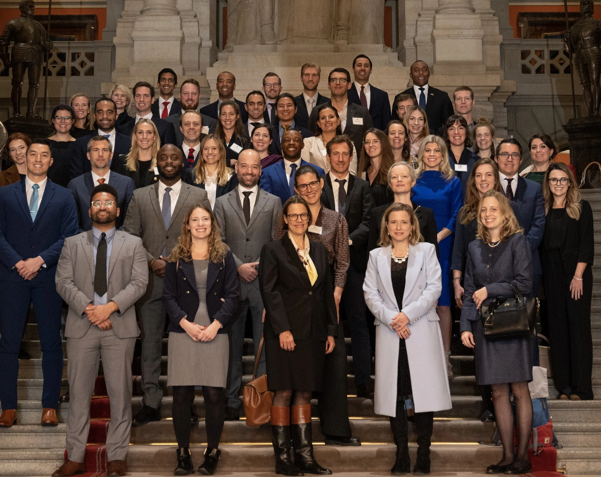 Group of diverse people in business attire standing on steps inside a grand building with statues and ornate architecture.