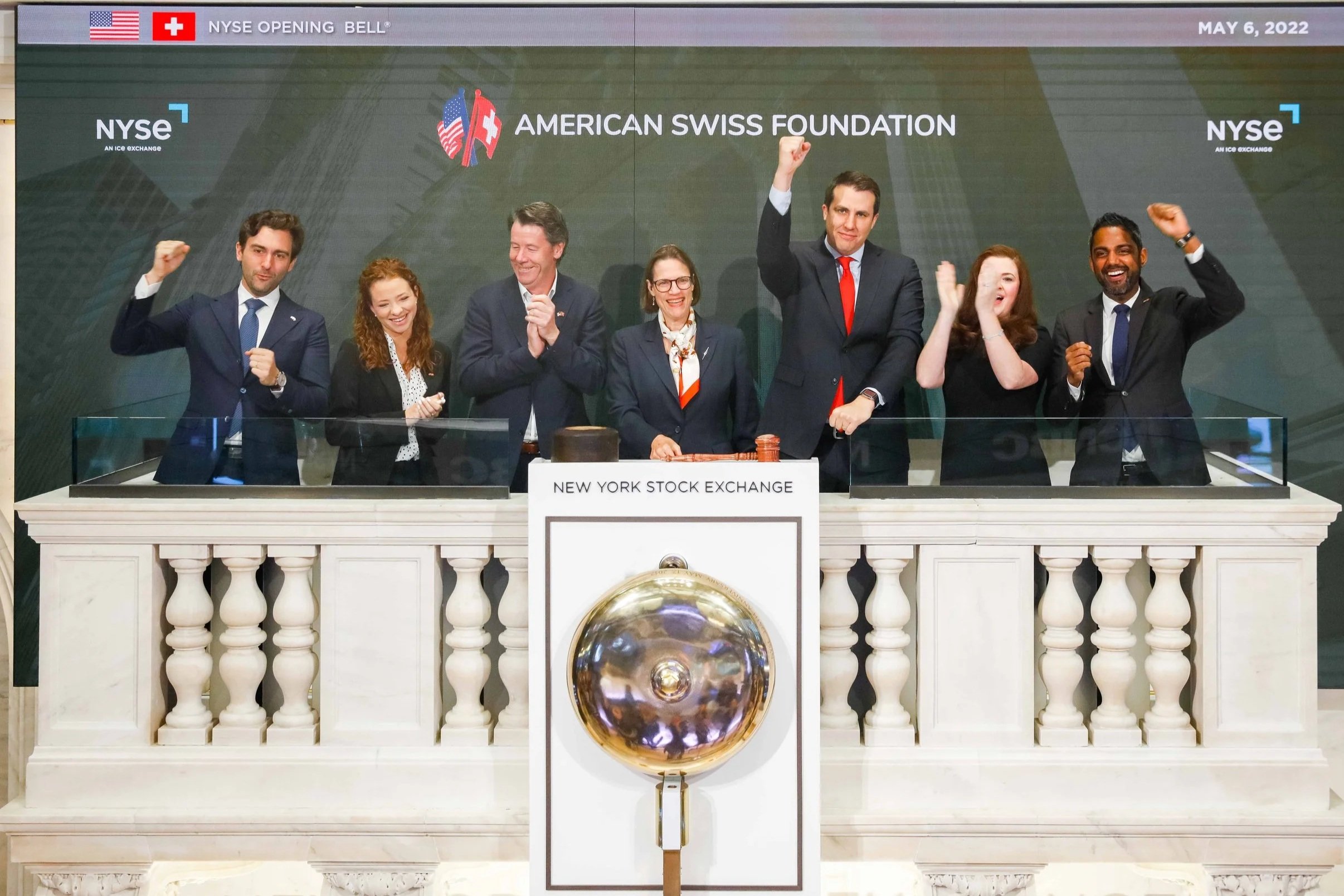 Seven people celebrating at the New York Stock Exchange balcony during the NYSE opening bell on May 6, 2022. They are dressed in business attire, smiling, and raising their fists in celebration. A large bell is in front of them, and a large display screen behind shows the event details and logos for NYSE and the American Swiss Foundation.