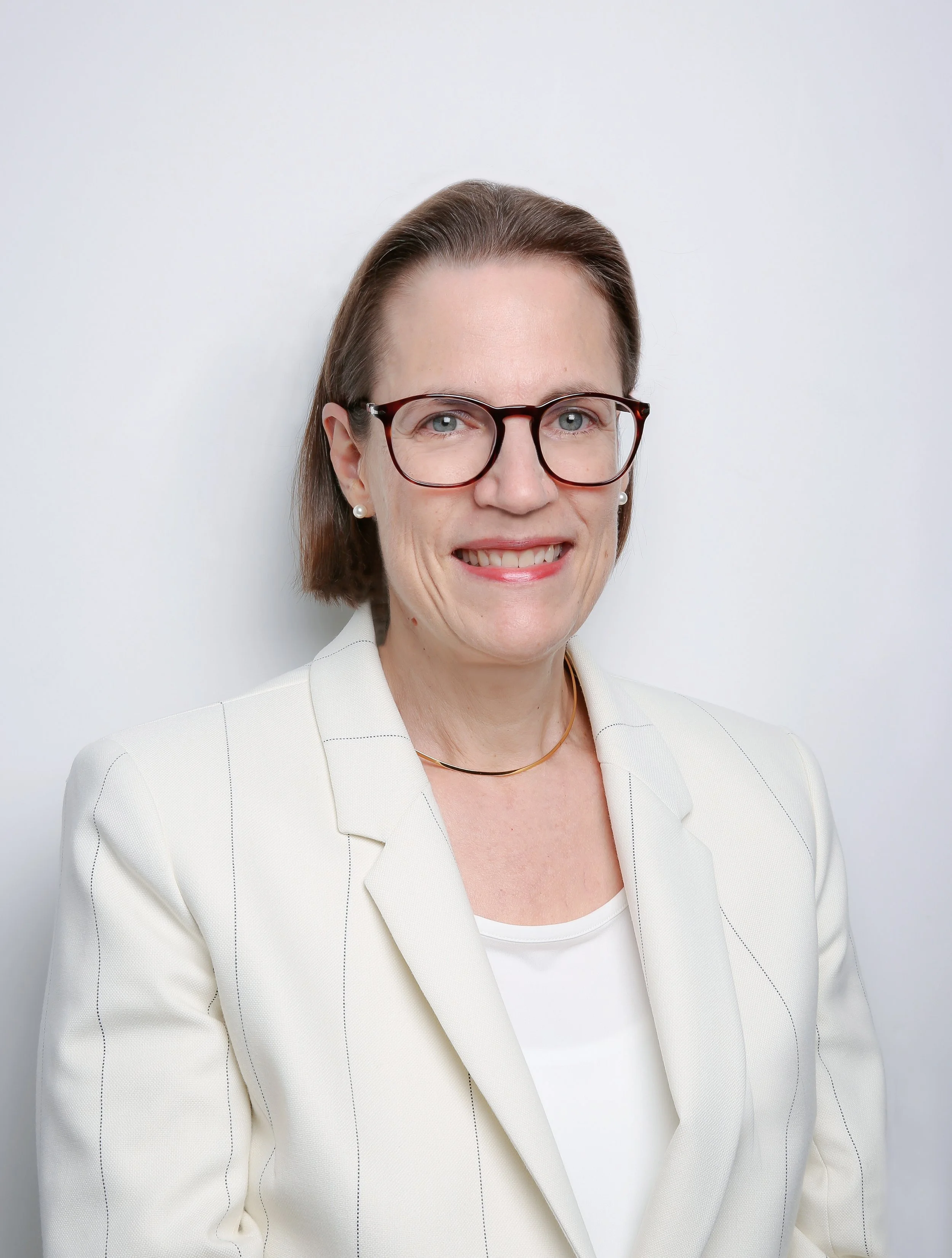 Headshot of a woman with short brown hair, wearing glasses, pearl earrings, a white blazer, and a gold necklace, smiling against a white background.