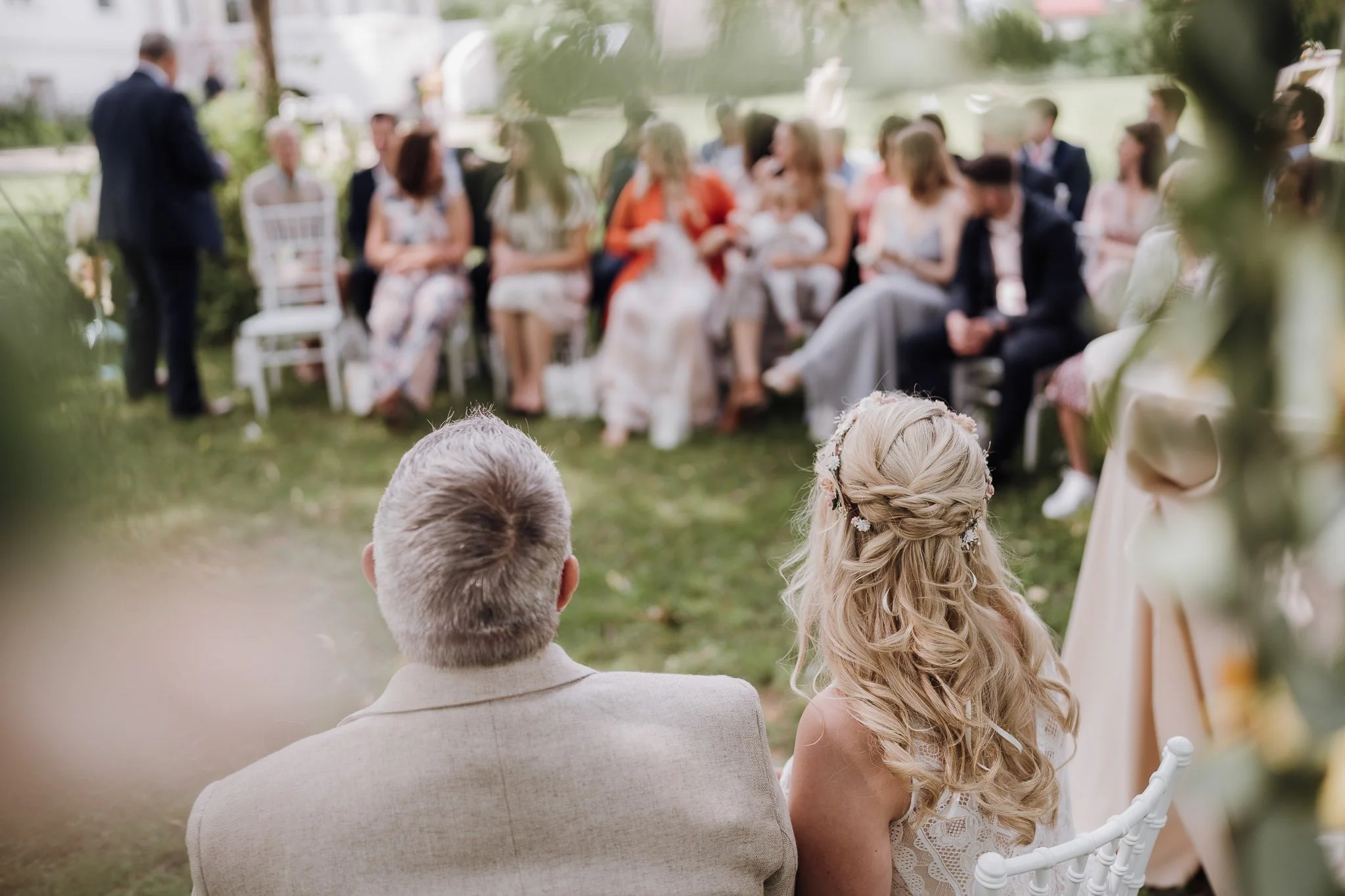 Im Vordergrund sitzendes mit dem Rücken zur Kamera gewandtes Hochzeitspaar, dass zu den Gästen schaut, während einer freien Trauung im engesten Familienkreis