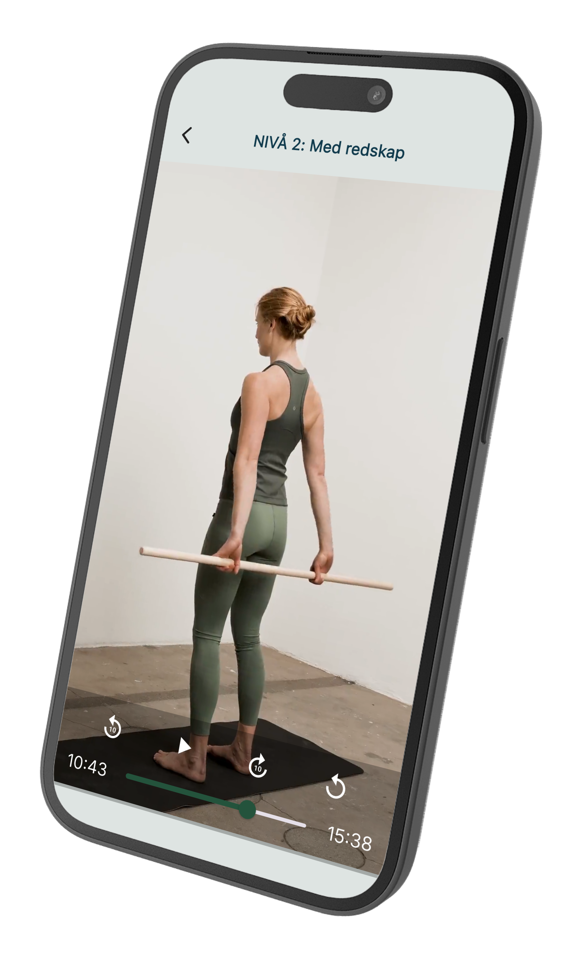 En kvinna står på en träningsmatta inomhus, håller ett långt trästickor bakom ryggen, klädd i träningskläder, och tittar nedåt.