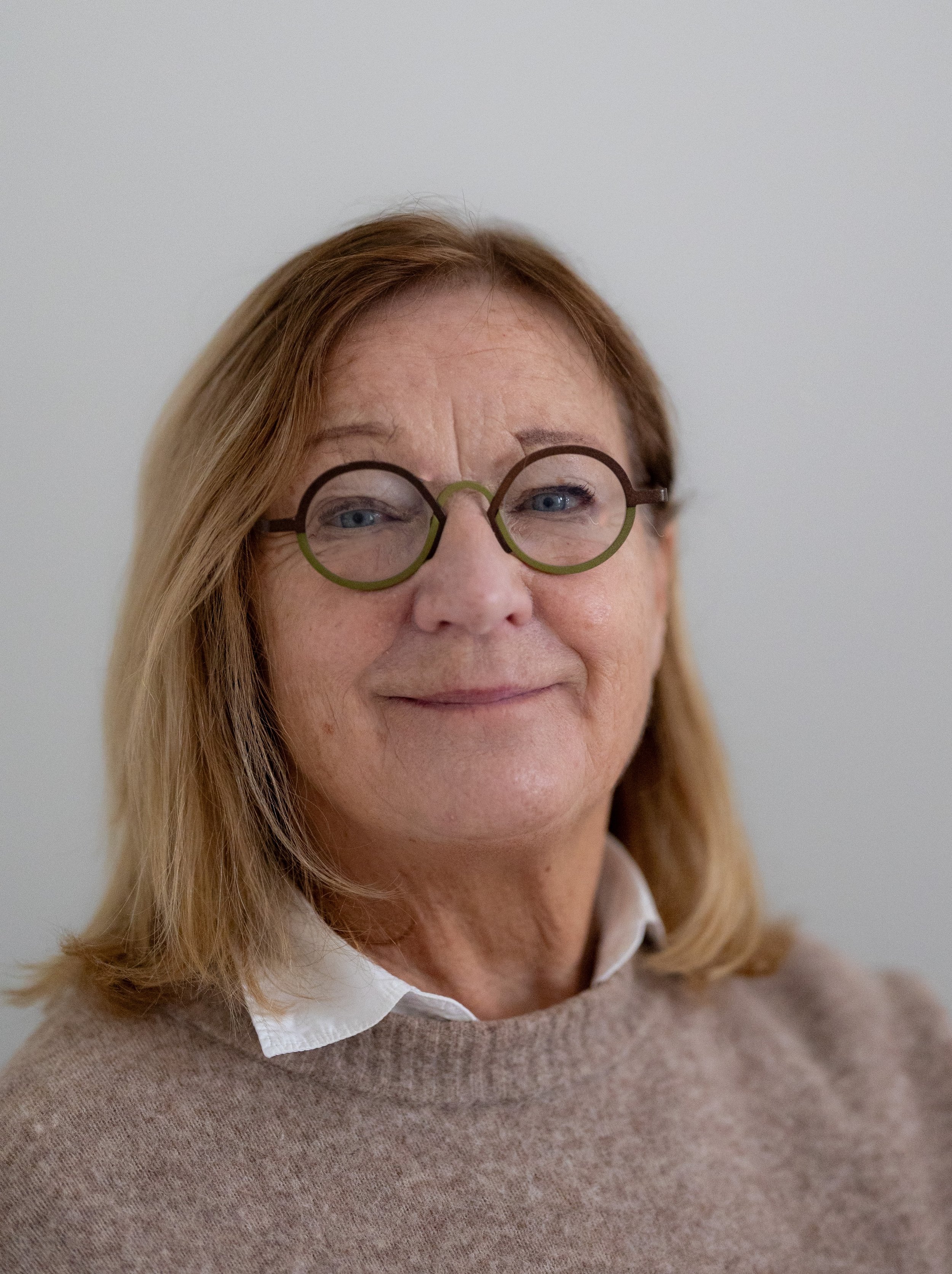 En kvinna med glasögon och ljusbrunt hår, ler lätt, står mot en vit vägg.
