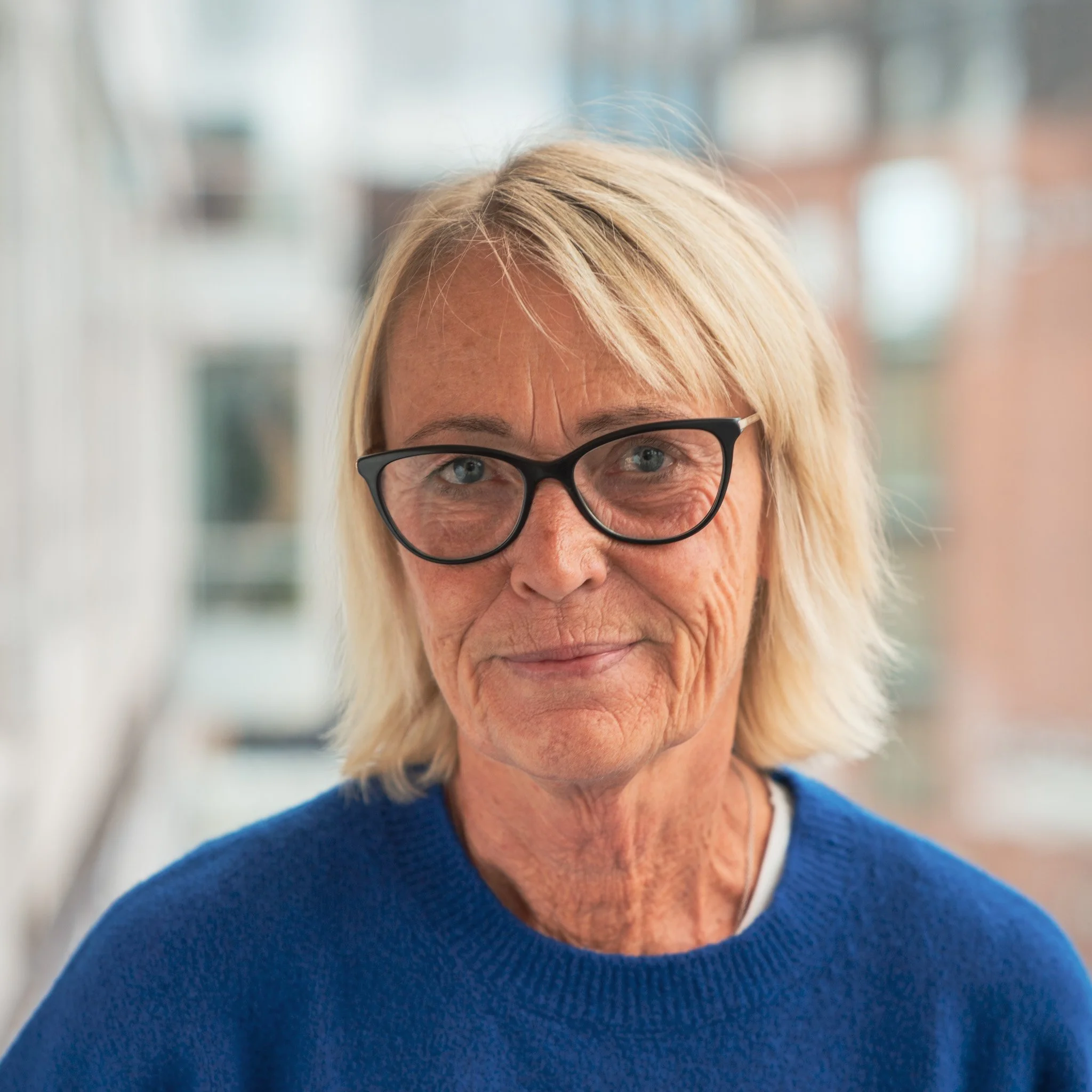 Porträtt av en äldre kvinna med blont hår, glasögon och en blå tröja, stående inomhus med en fönsterbygd i bakgrunden, som tränar via osteoporosgymmet