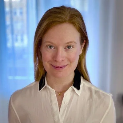 En kvinna med brunt hår och vitt skjorta, genom ett fönster i bakgrunden.