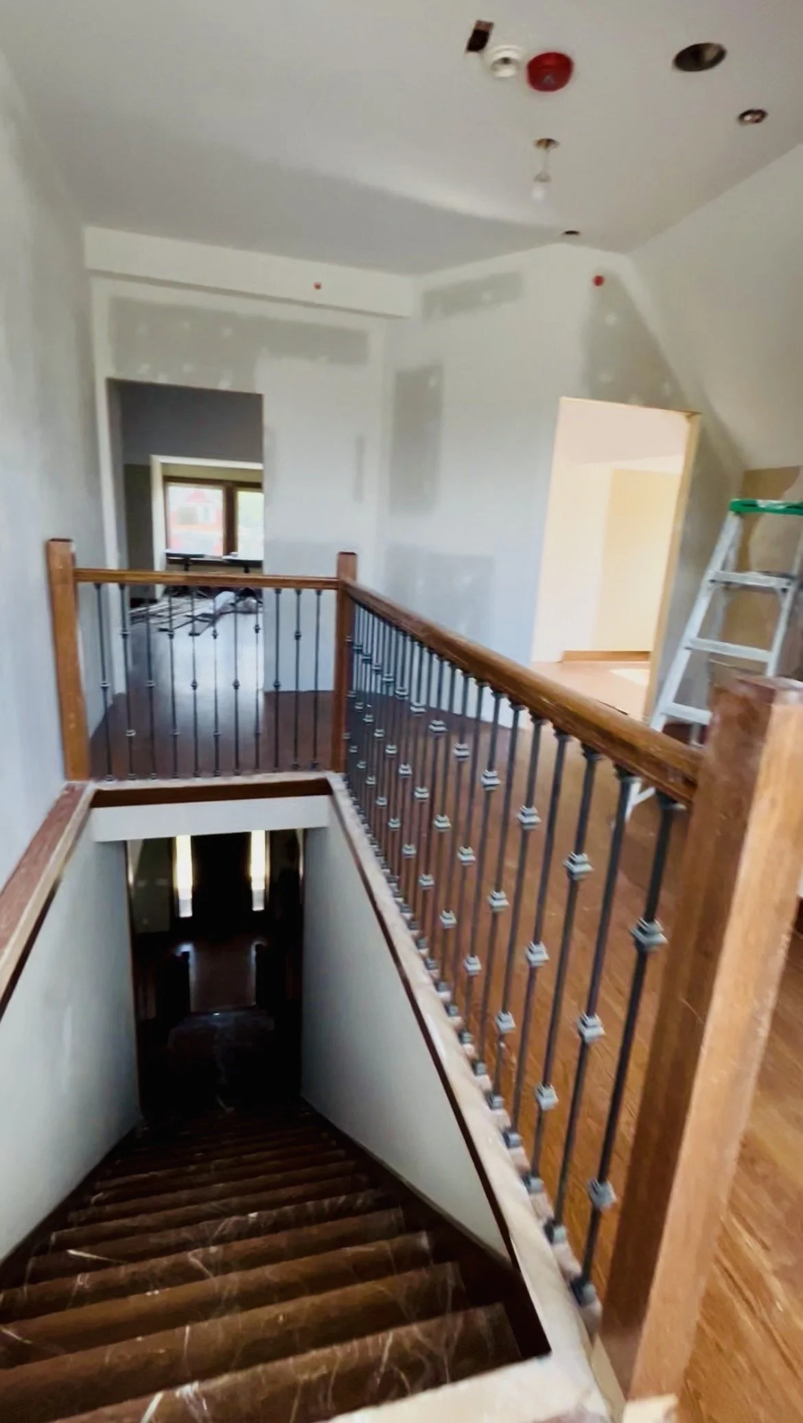 Interior view of a house under renovation showing a wooden staircase, a half-finished white wall, and a small upstairs landing area with a ladder and exposed ceiling fixtures.