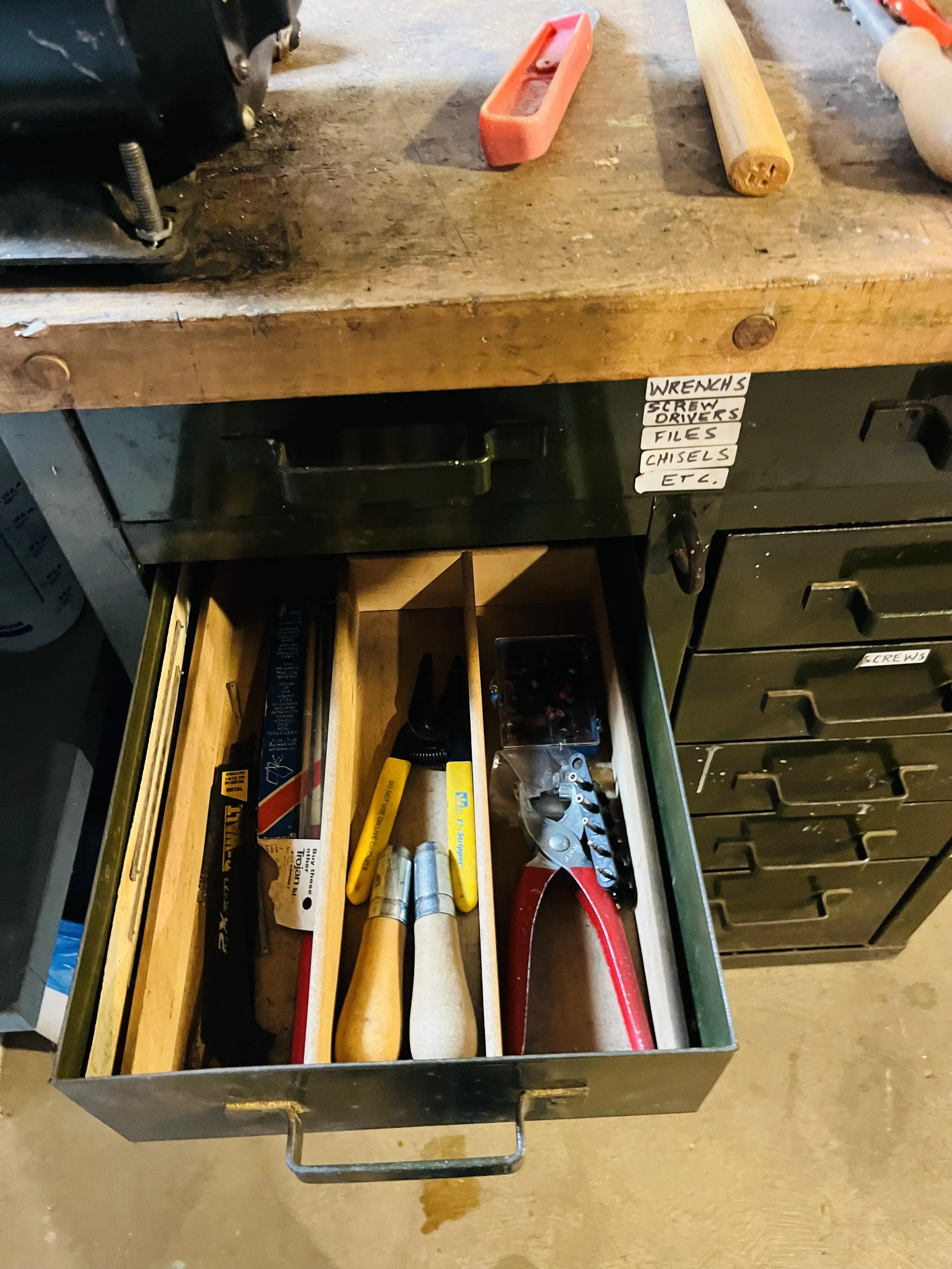 An open drawer in a workshop containing screwdrivers, pliers, files, and other hand tools, with a wooden workbench above and shelving labels for tools.