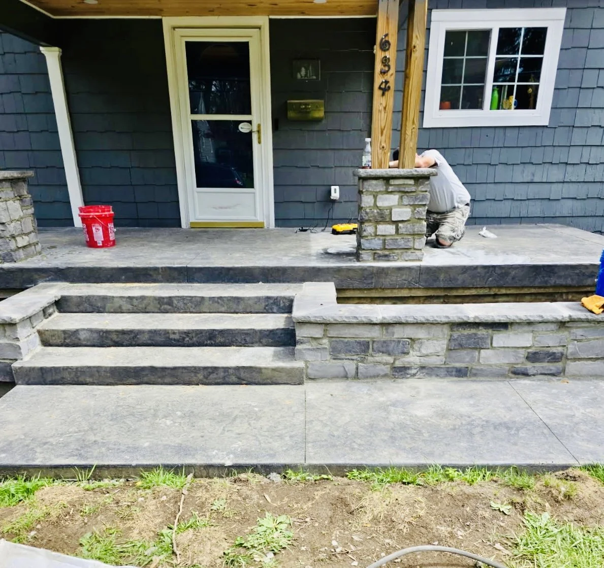 A house with a gray exterior and white trim shows a porch with stone steps. A person is kneeling, working on the porch near a brick column with the house number 6934. There is a red bucket on the porch and various tools and bottles nearby.