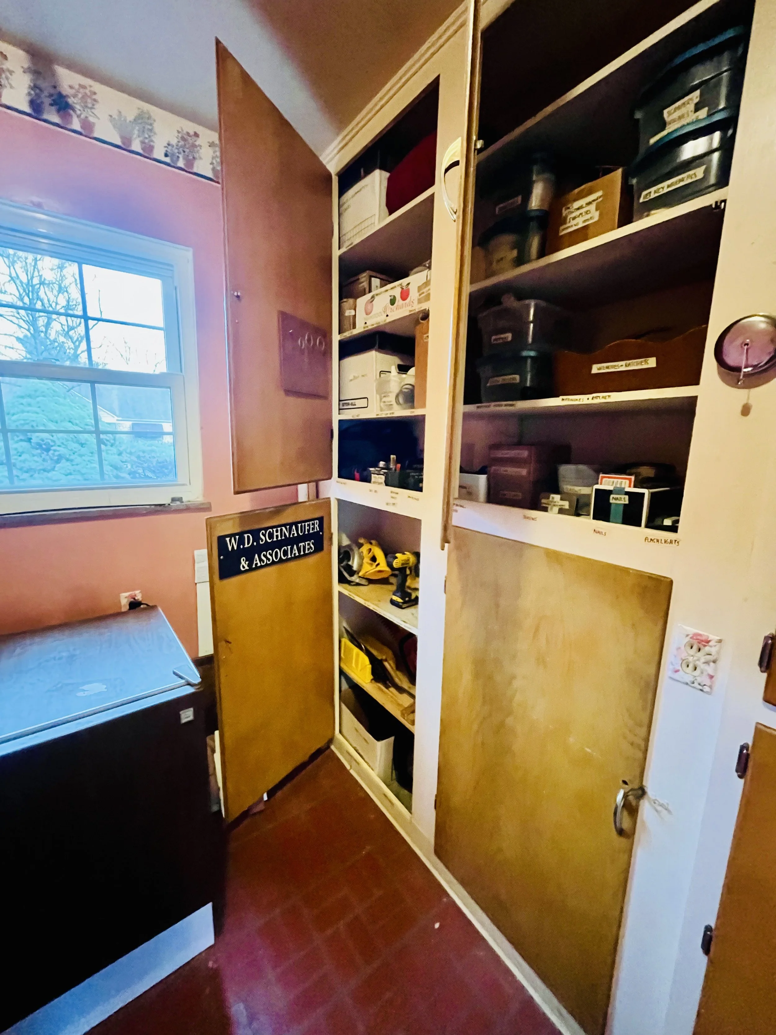 A pantry with open shelves holding storage bins, tools, and boxes, next to a window with a view of trees outside.