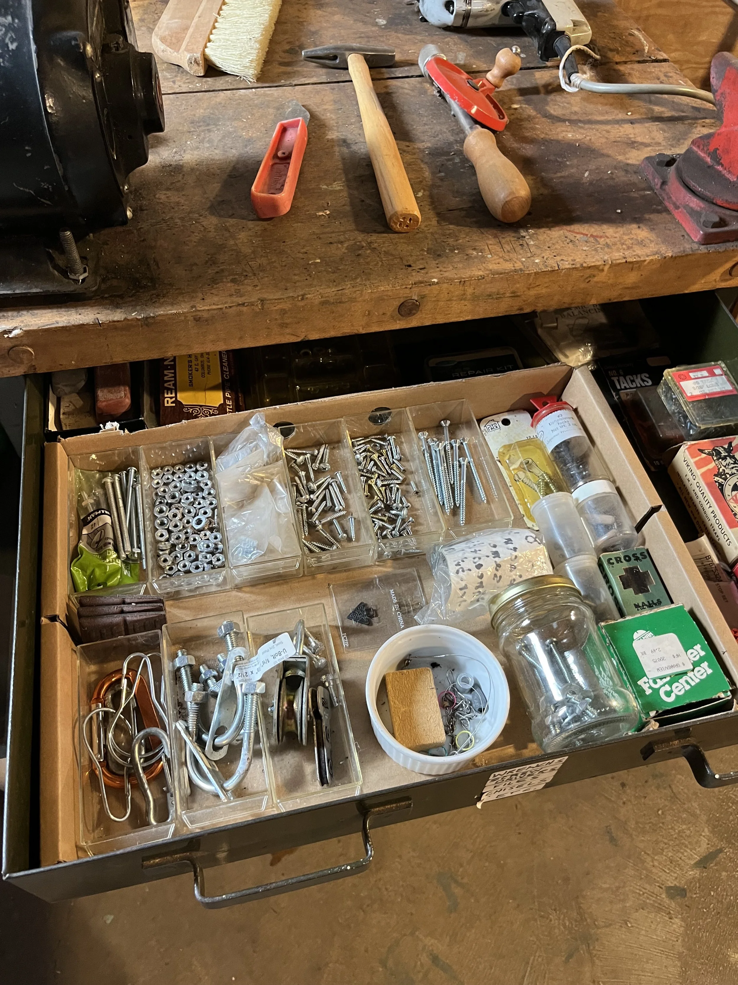 Open toolbox drawer with various small hardware parts including screws, nails, hooks, and other fasteners, along with a small container holding assorted items and a green box labeled 'First Aid Center.'