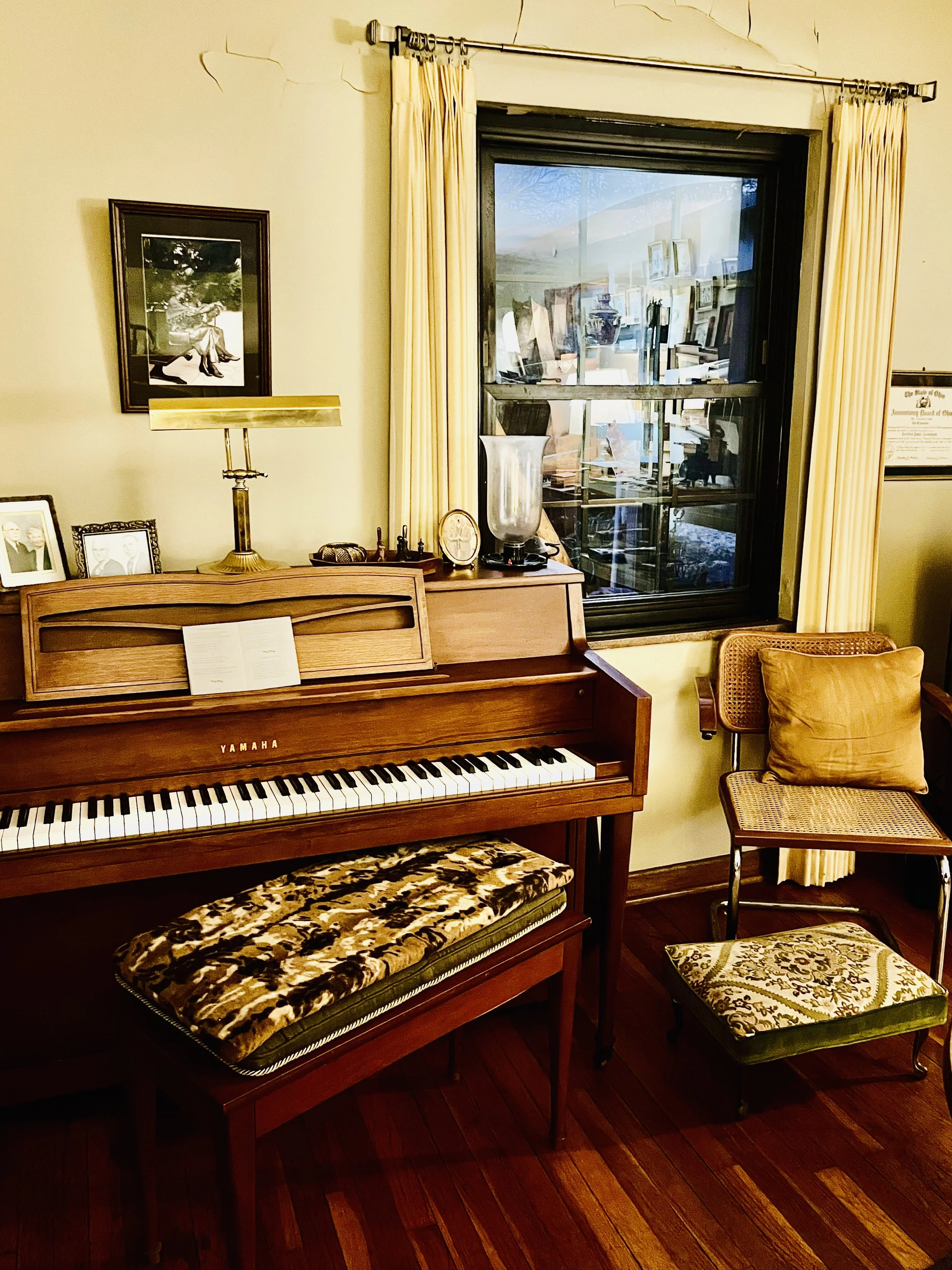 A cozy corner with a Yamaha upright piano, a bench with a patterned cushion, and a wooden chair with a yellow pillow. There are framed photographs and a lamp on top of the piano, and a large window with yellow curtains showing a reflection of a room 