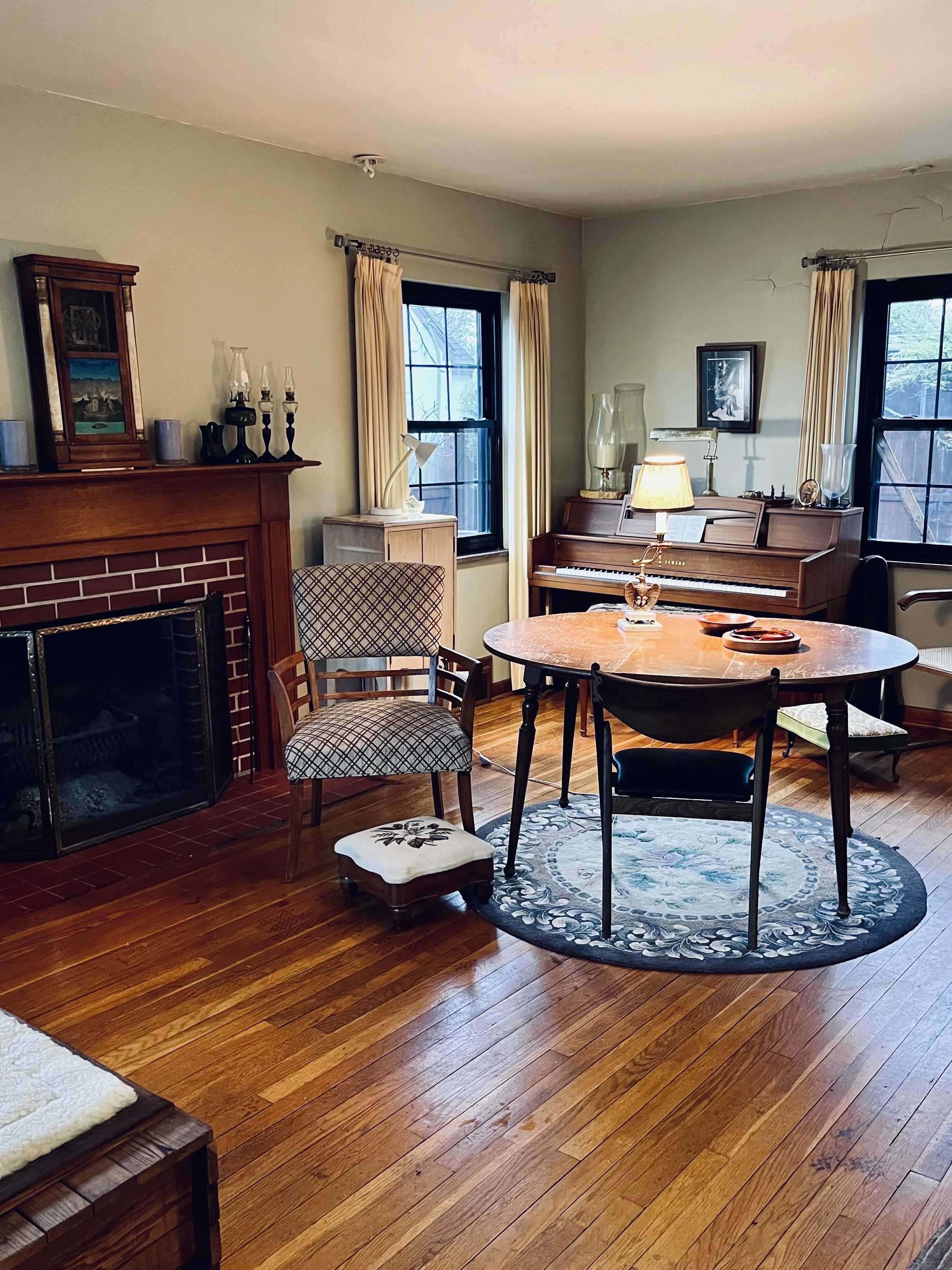 A cozy living room with hardwood floors, a round rug, a wooden dining table, various chairs, a piano, fireplace, and large windows with curtains.