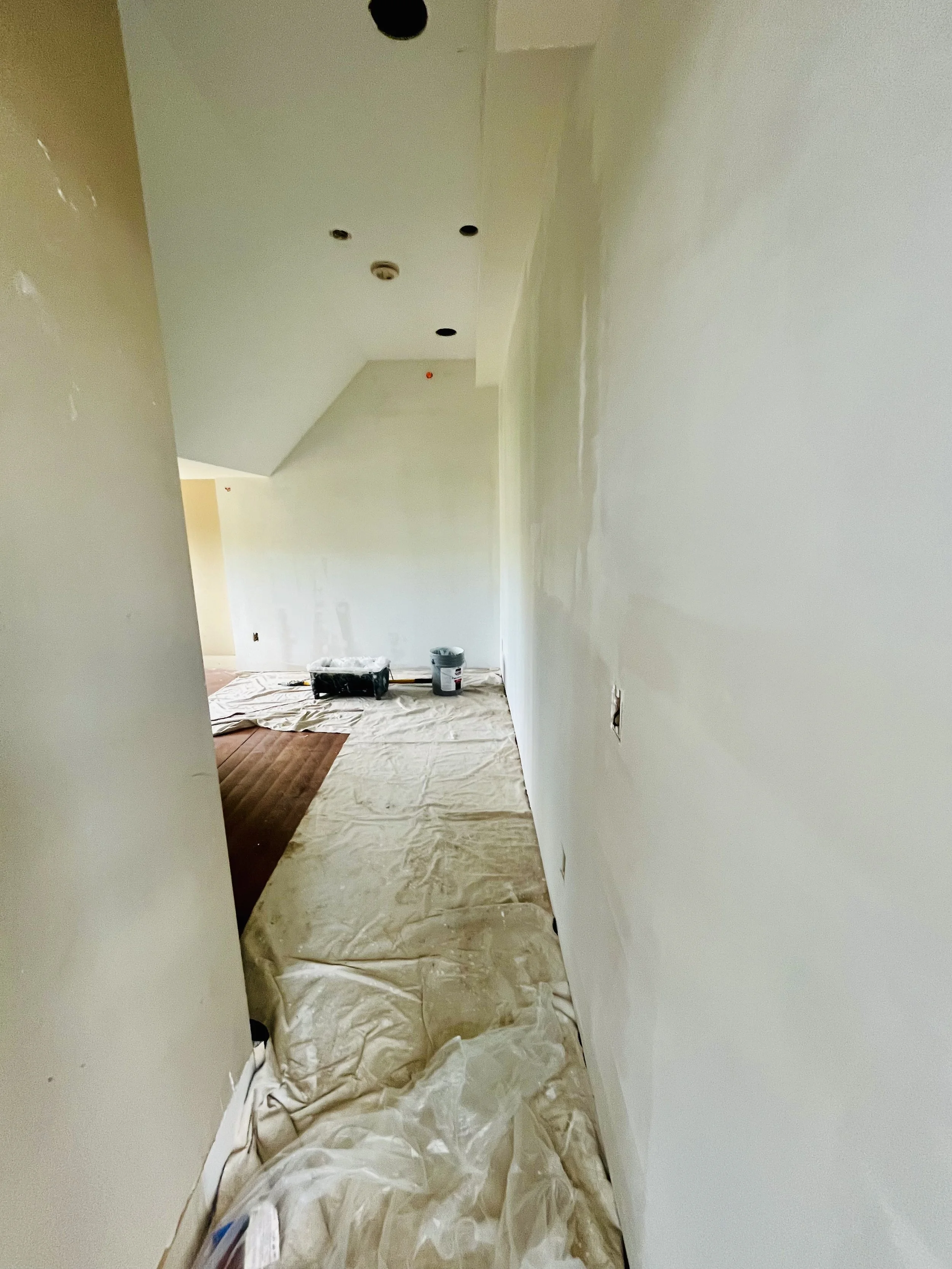Interior of a house under construction or renovation with partially painted walls, ceiling lights not yet installed, and protective plastic covering the floor.