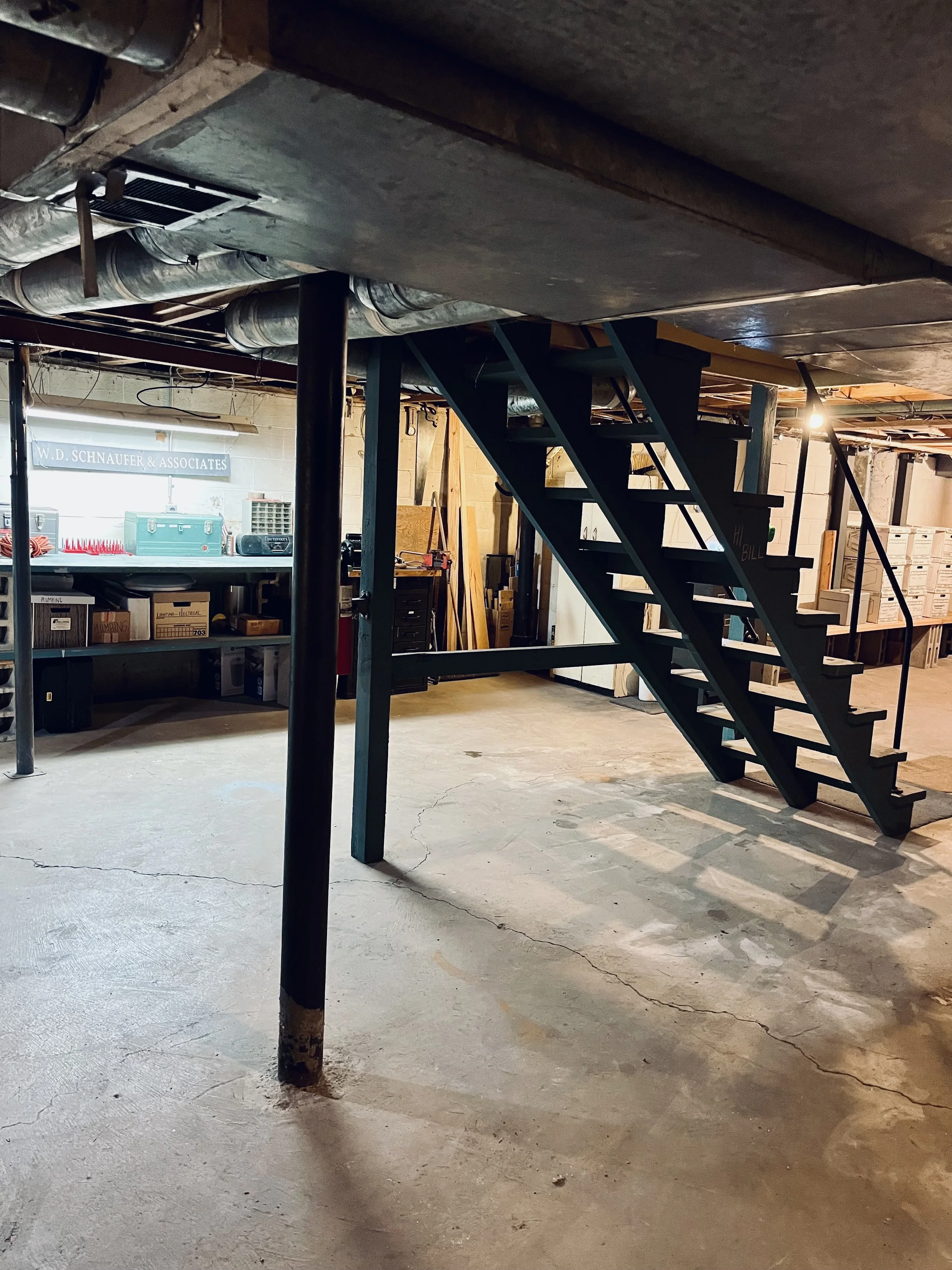 Basement with concrete floor, black metal staircase, exposed pipes and ductwork, and a workbench with tools and boxes.