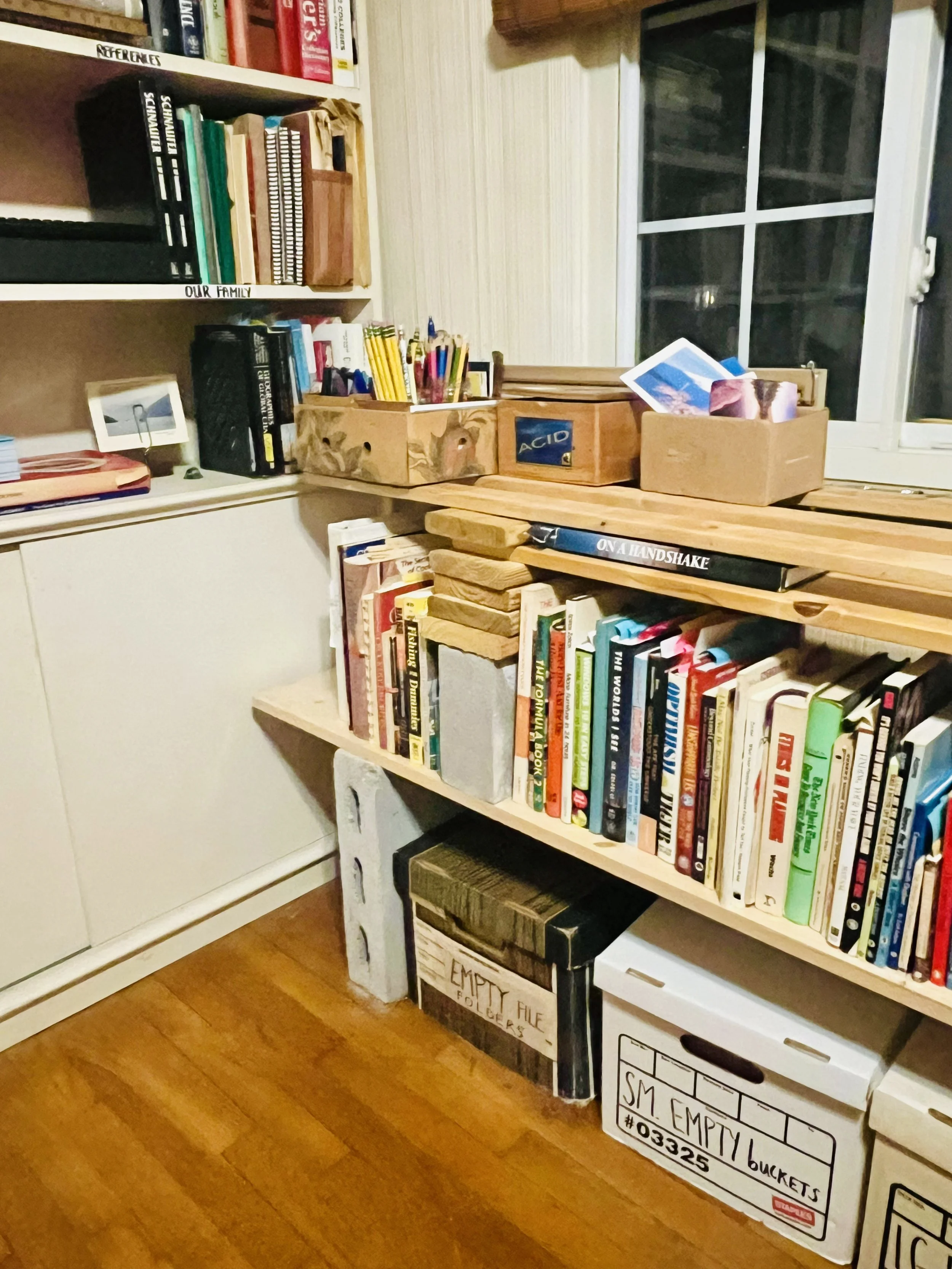 A cluttered room with bookshelves, books, and boxes, including a white box labeled 'SM EMPTY BUCKETS,' a cardboard box marked 'EMPTY FILE FOLDER,' and a brown wooden box with pens and papers on a wooden table near a window.