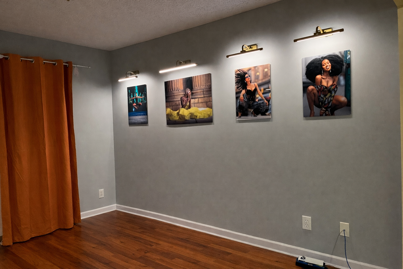 Gallery wall with four framed photographs of diverse women in various poses, illuminated by picture lights, in a room with a gray wall, hardwood floor, and orange curtains.