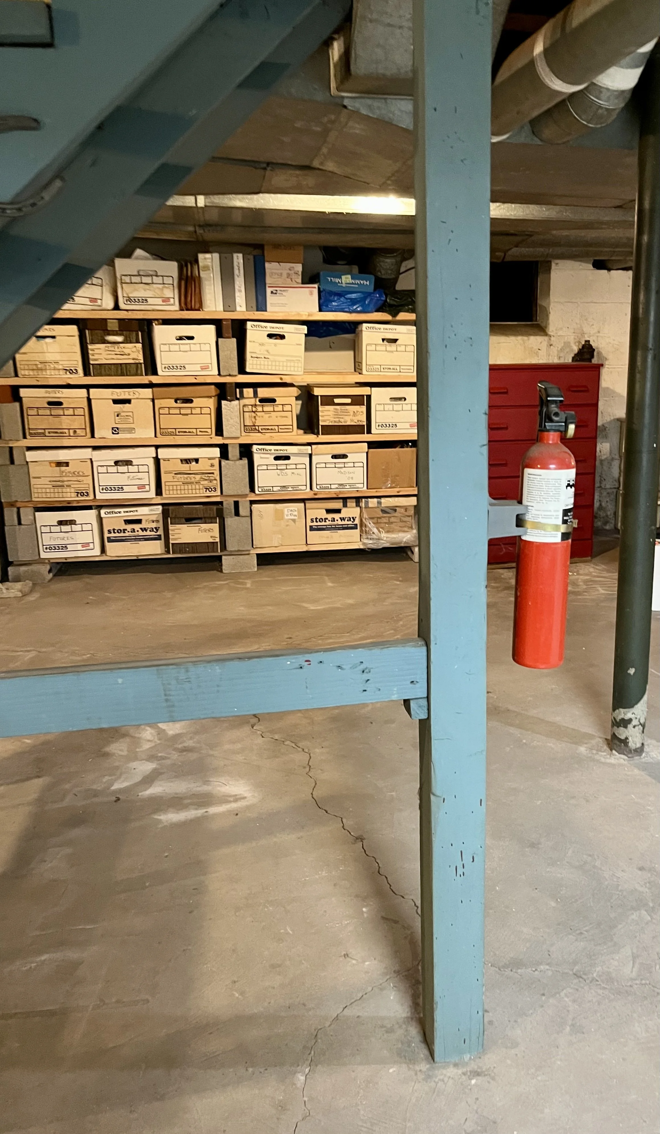 Storage area with a wooden shelf filled with labeled cardboard boxes, a red toolbox, a fire extinguisher mounted on a post, and cracks on the concrete floor.