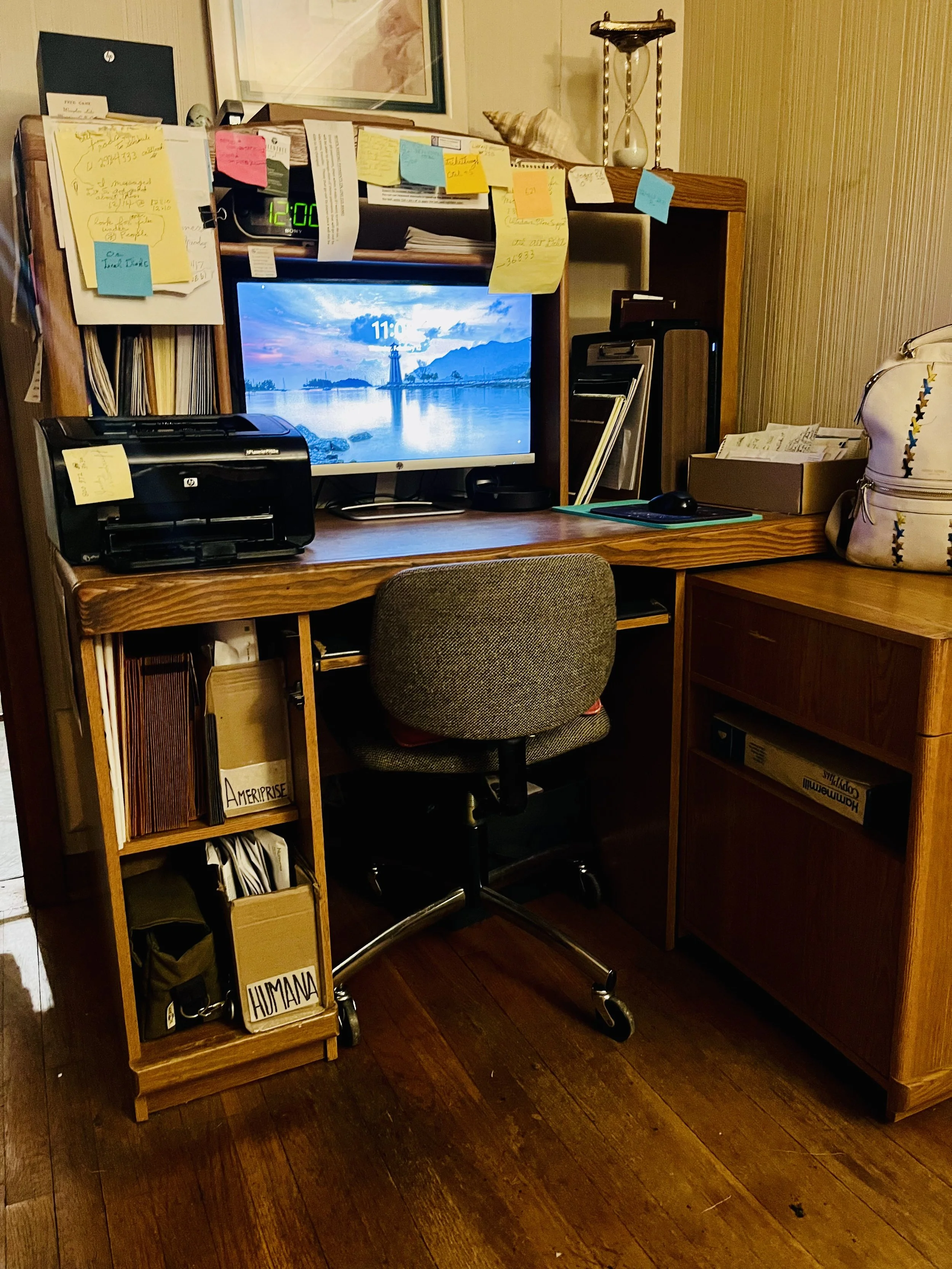 A cluttered wooden desk with an abundance of sticky notes, papers, and folders. A computer monitor displays a scenic landscape with clouds and mountains. There is a printer on the left and a small bookshelf on the left with folders labeled "Amerprise