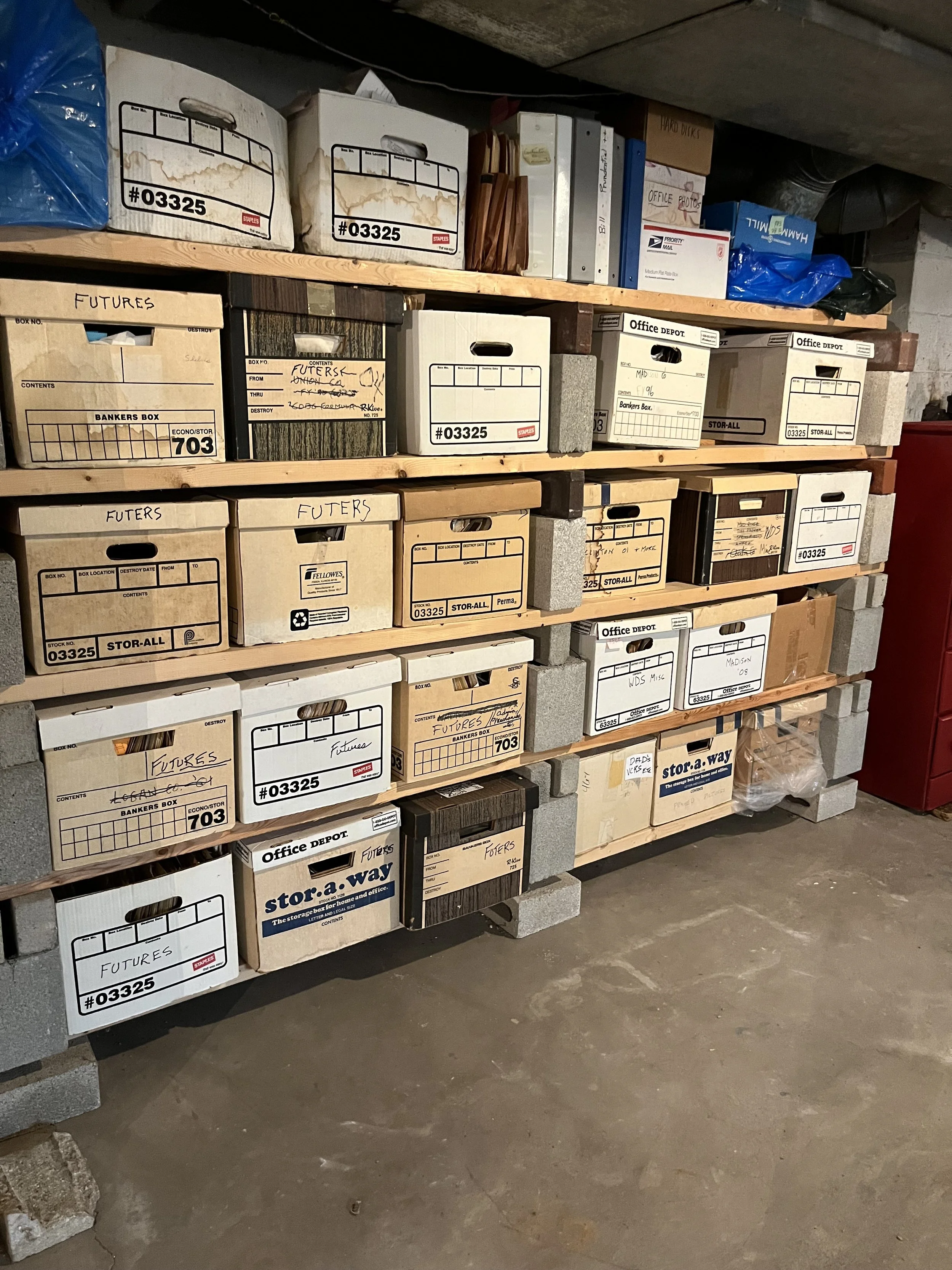 Shelves with cardboard and plastic bins filled with files, documents, and office supplies in a storage room.