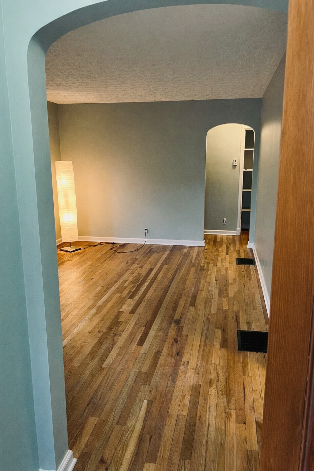 Empty living room with hardwood floors, a tall floor lamp, and a small corner space with built-in shelves.