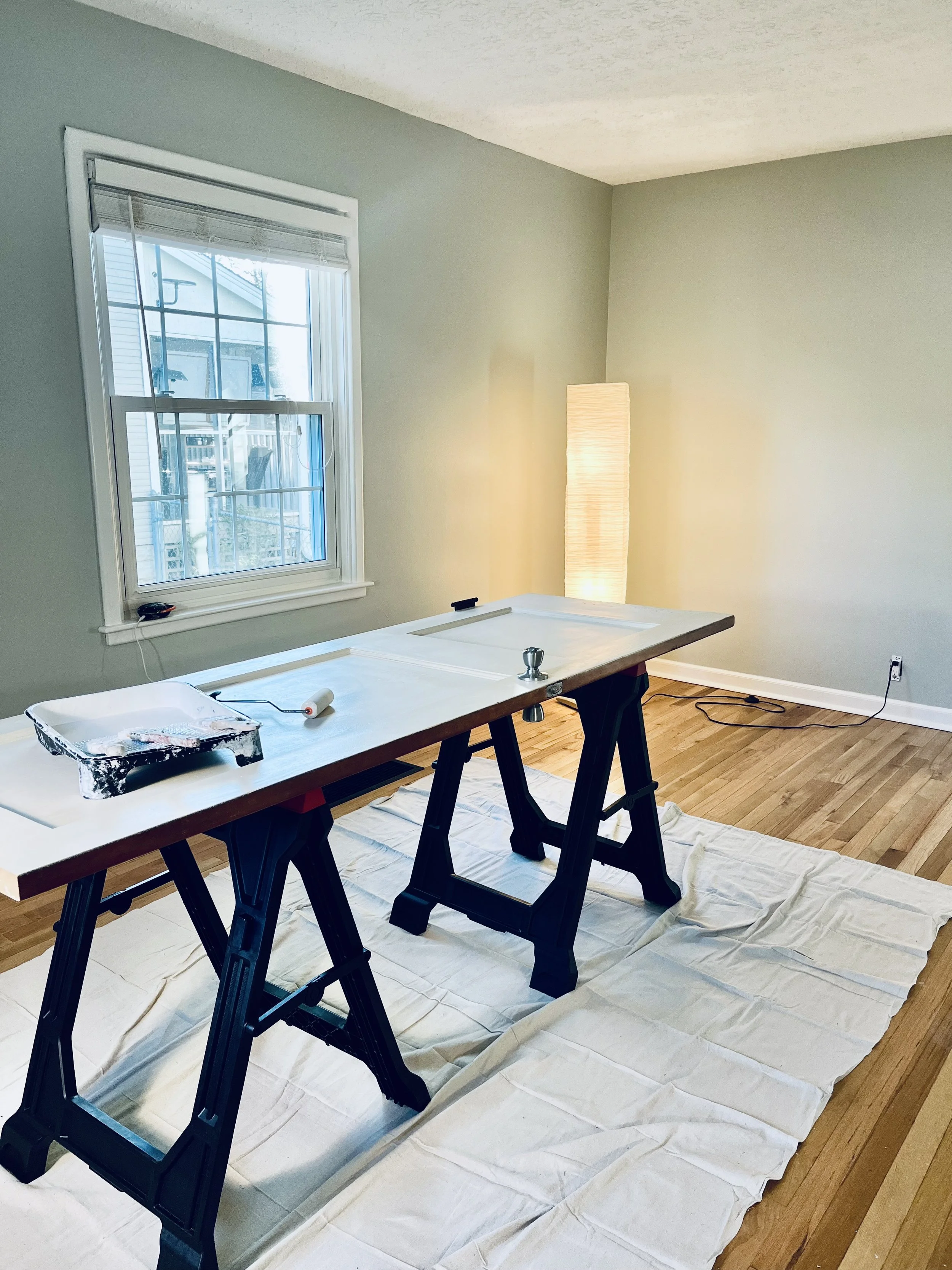 Room with a window, a tall floor lamp, and a large wooden table on sawhorses covered with a drop cloth, with painting tools and roller on the table.