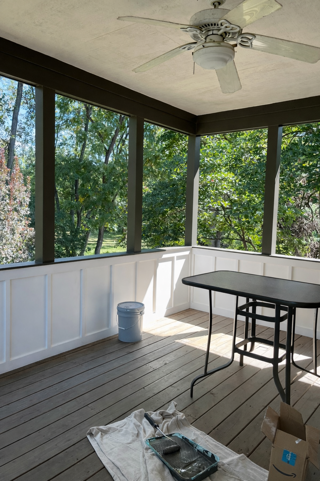 A screened porch with a ceiling fan, a plastic bucket, and a table, with trees visible through the screening. Painting supplies and a cardboard box are on the wooden floor.