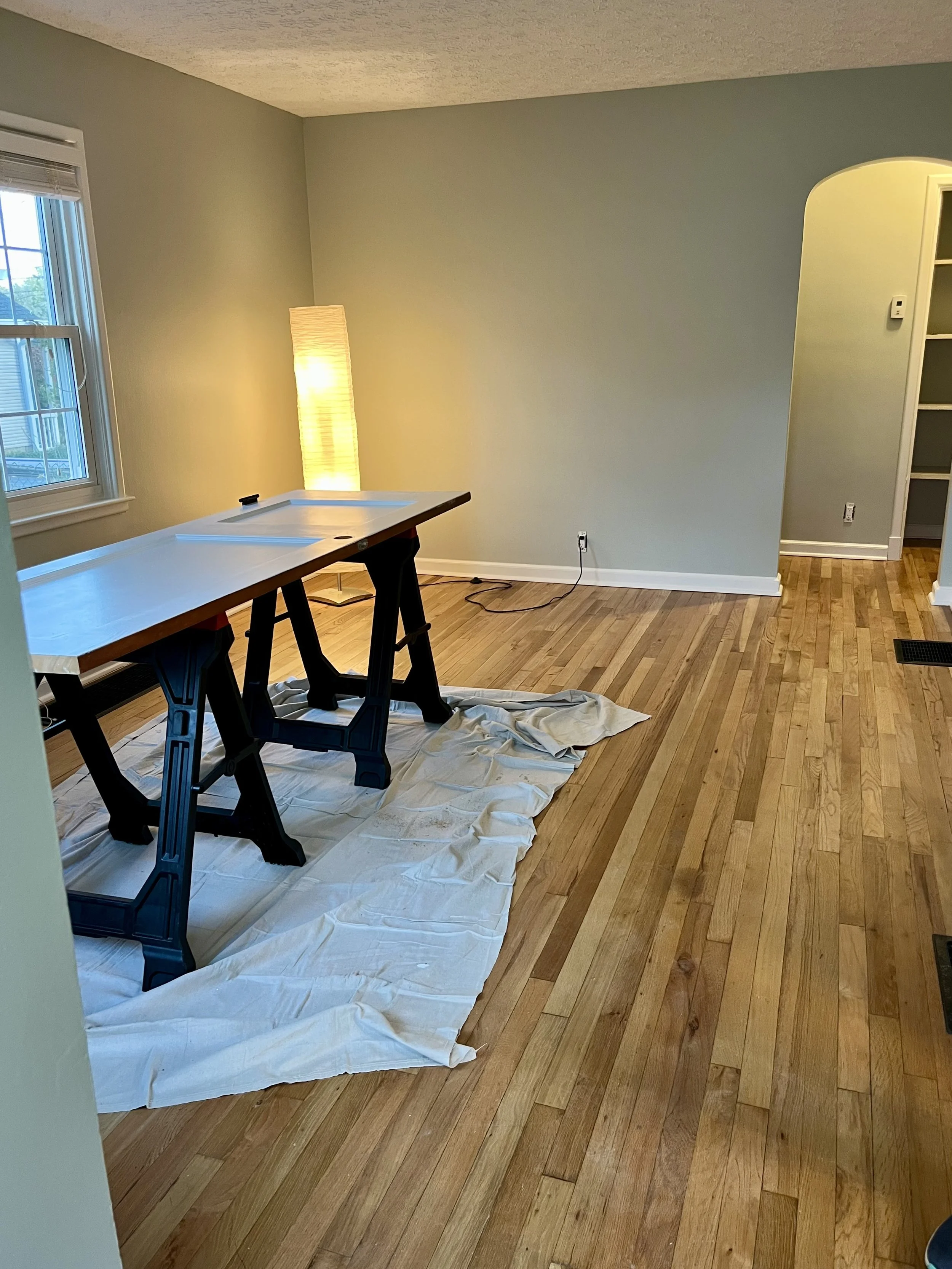 Interior of a room with hardwood flooring, a window on the left, a tall floor lamp emitting warm light, and a work table covered with a white cloth, possibly for painting or renovation, with an electrical cord plugged into an outlet on the back wall.