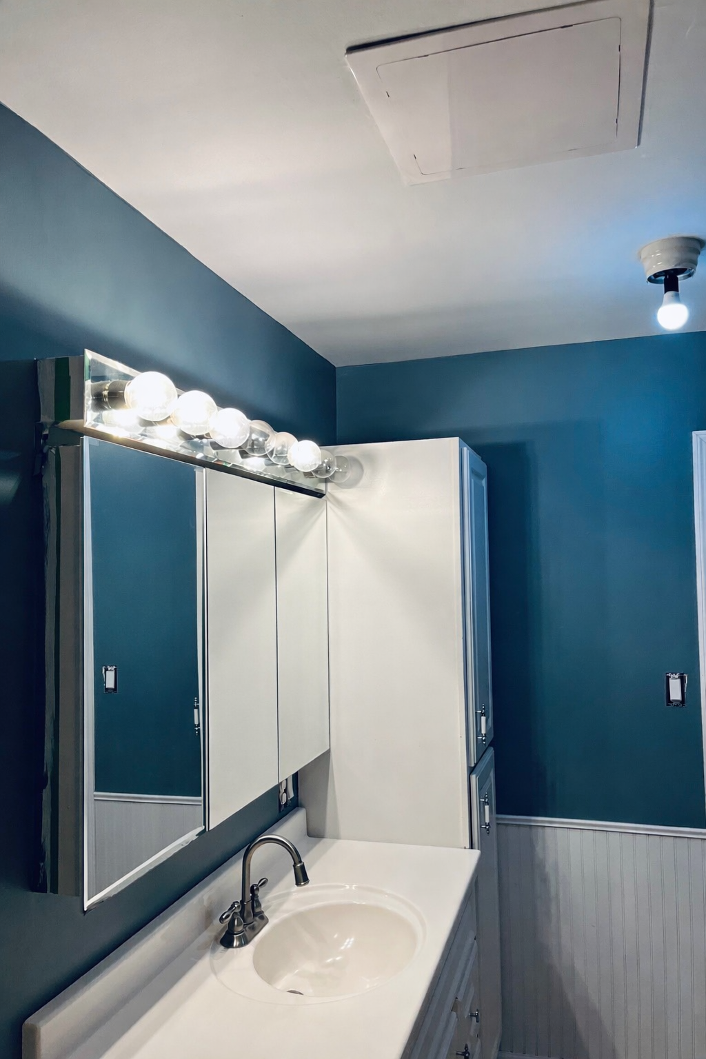 A bathroom with a white vanity, a mirror with lighting fixtures, a blue wall, and a ceiling vent.