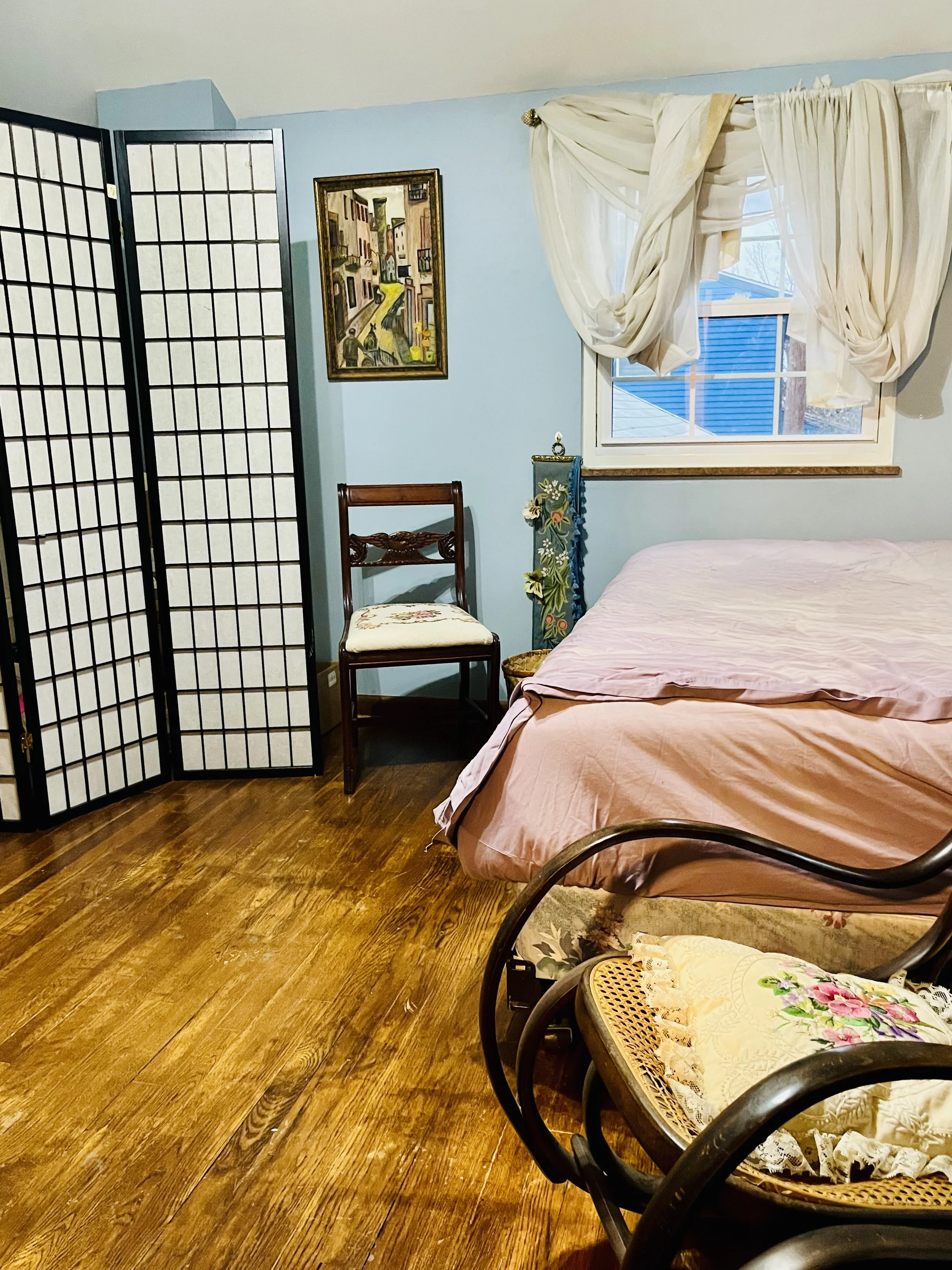 A cozy bedroom with light blue walls, a window with cream curtains, a bed with pink sheets, a wooden chair with a floral cushion, a decorative screen, an artwork, and a small floral stand.