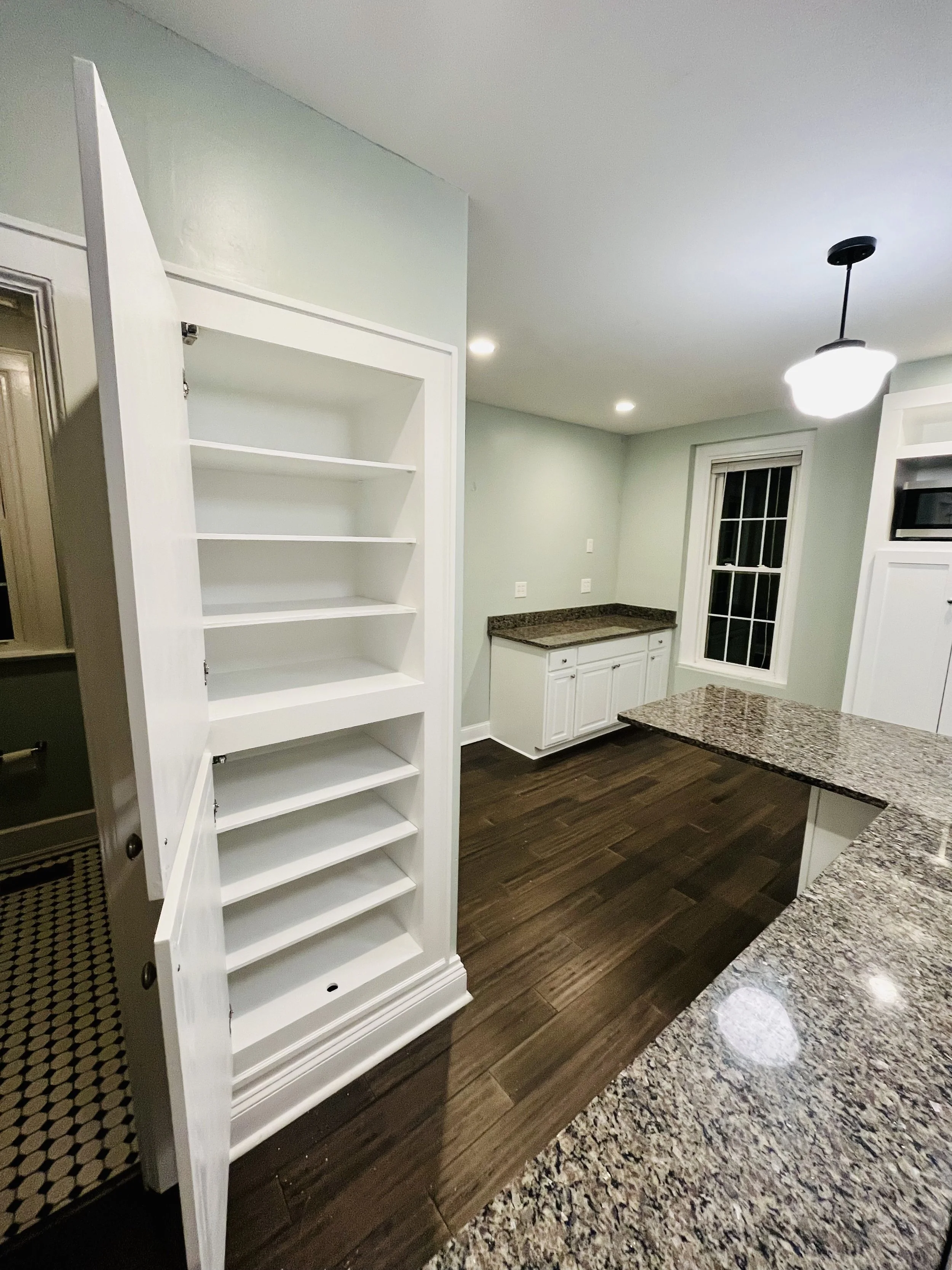 Spacious kitchen with granite countertops, white cabinets, a window, and a tall white cabinet with shelves.