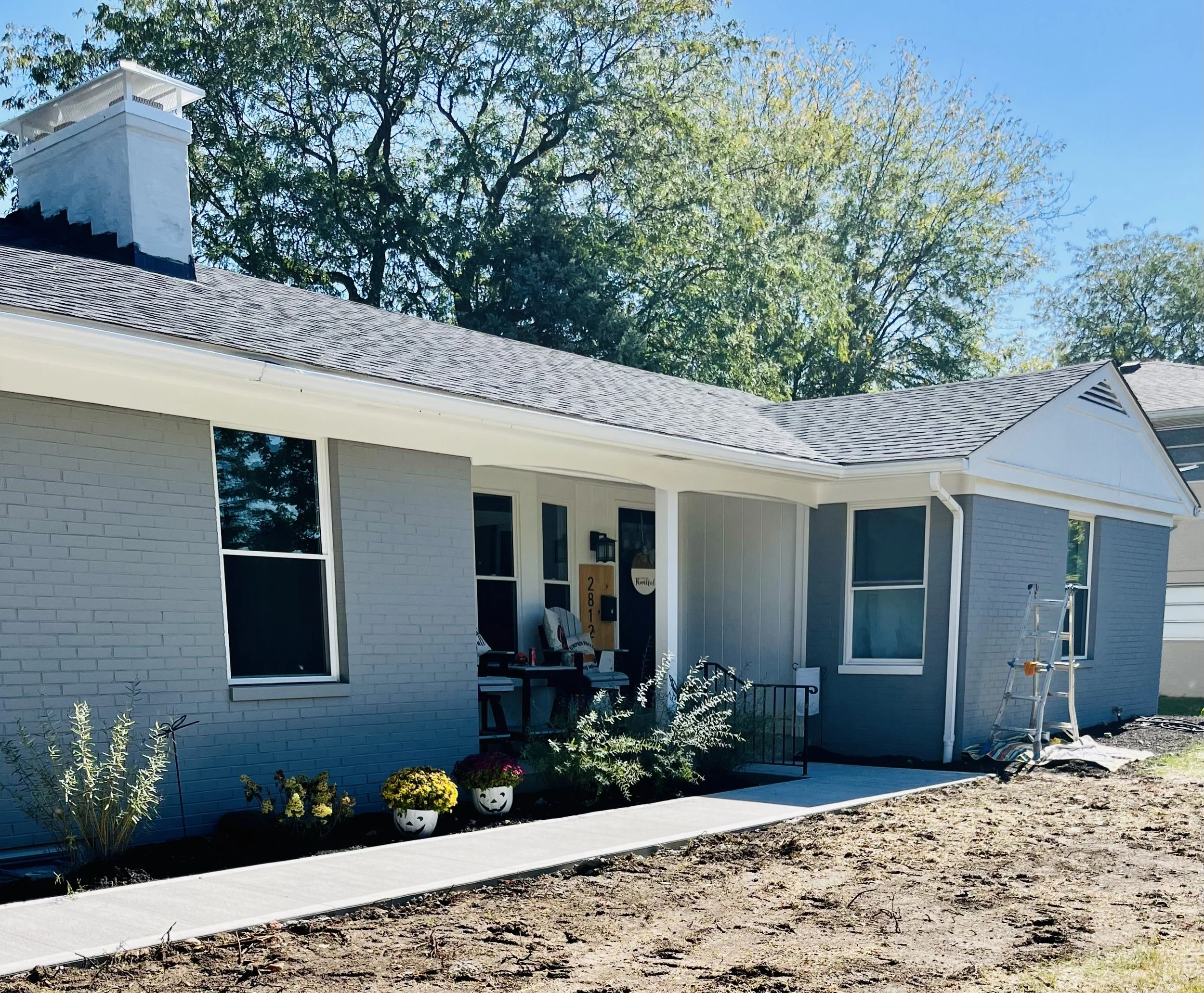 A newly renovated house with a light blue brick exterior, large windows, a front porch with a welcome sign, and small decorative pumpkins and flowers on the front steps. A ladder leans against the house, and the yard is being landscaped.