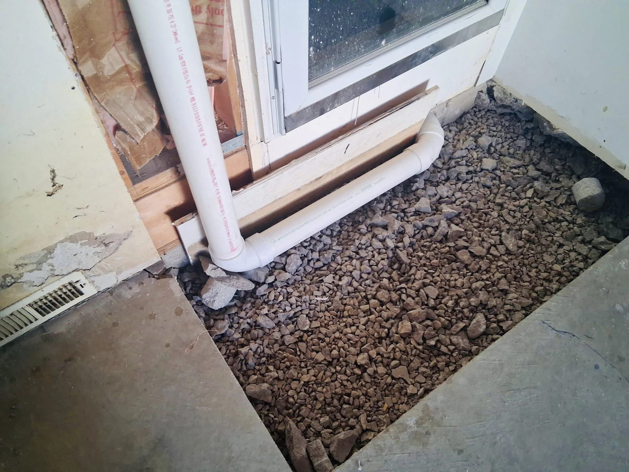 A construction site showing a floor with gravel and rocks, PVC pipes running along the base of a sliding glass door, and a vent on the floor near a wall.