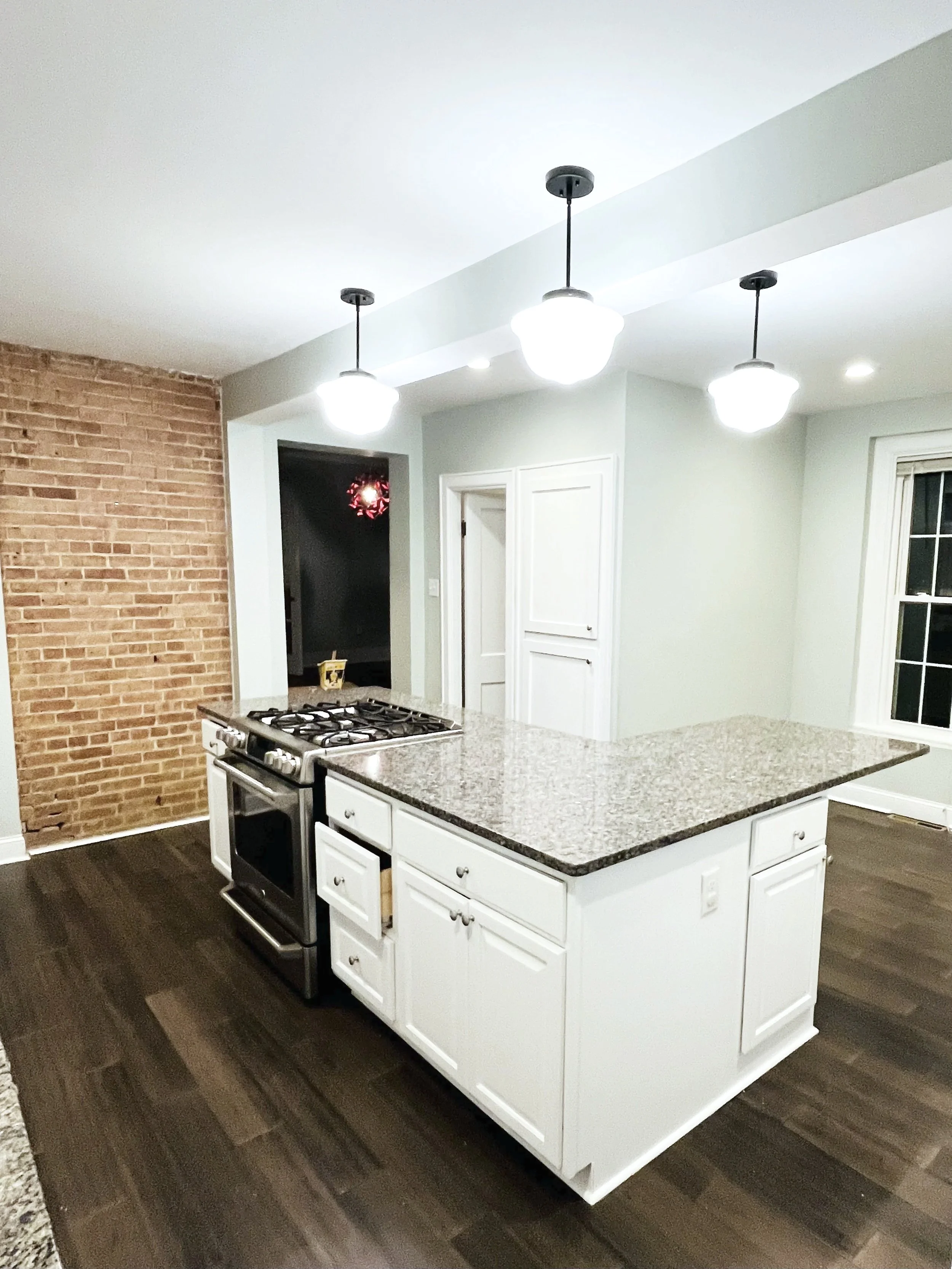 A modern kitchen with white cabinets, a granite countertop island, a stainless steel stove, and pendant lights hanging from the ceiling. A brick accent wall is visible on the left, and a window with white trim is on the right.