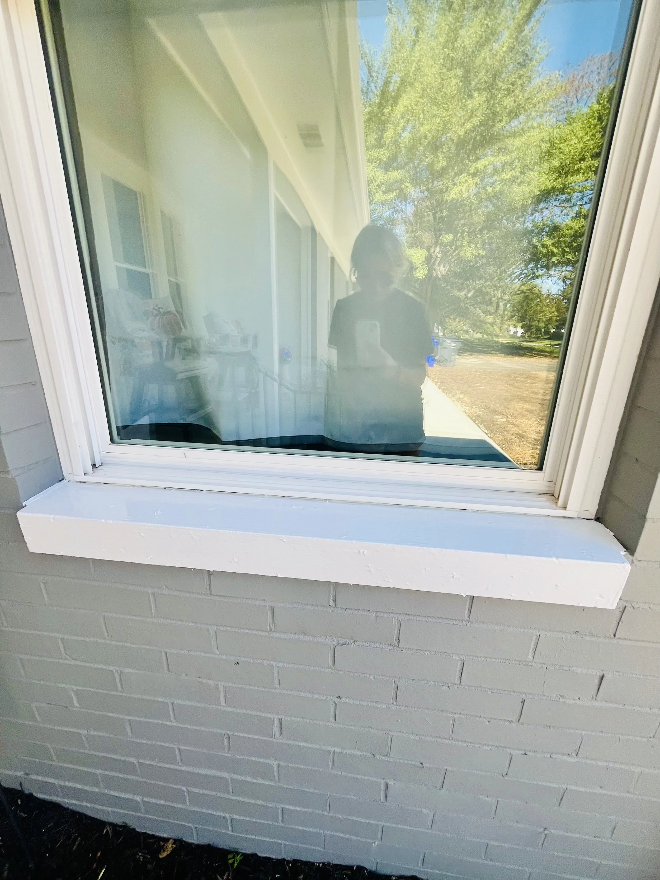 Reflection of a woman taking a photo in a window with a tree outside and a porch visible.