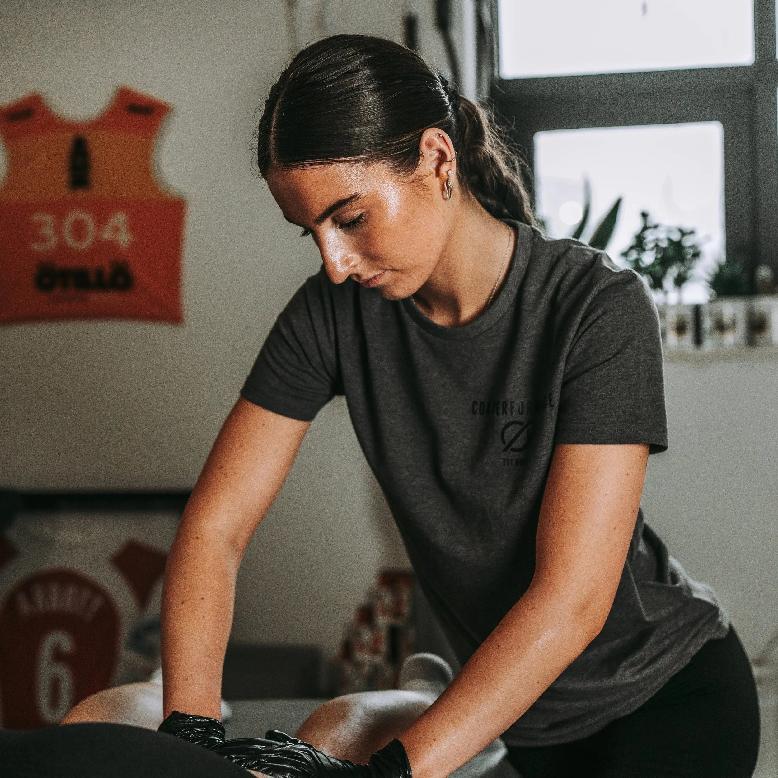 Young woman giving a massage in a room with sports jerseys on the wall and a window with plants.