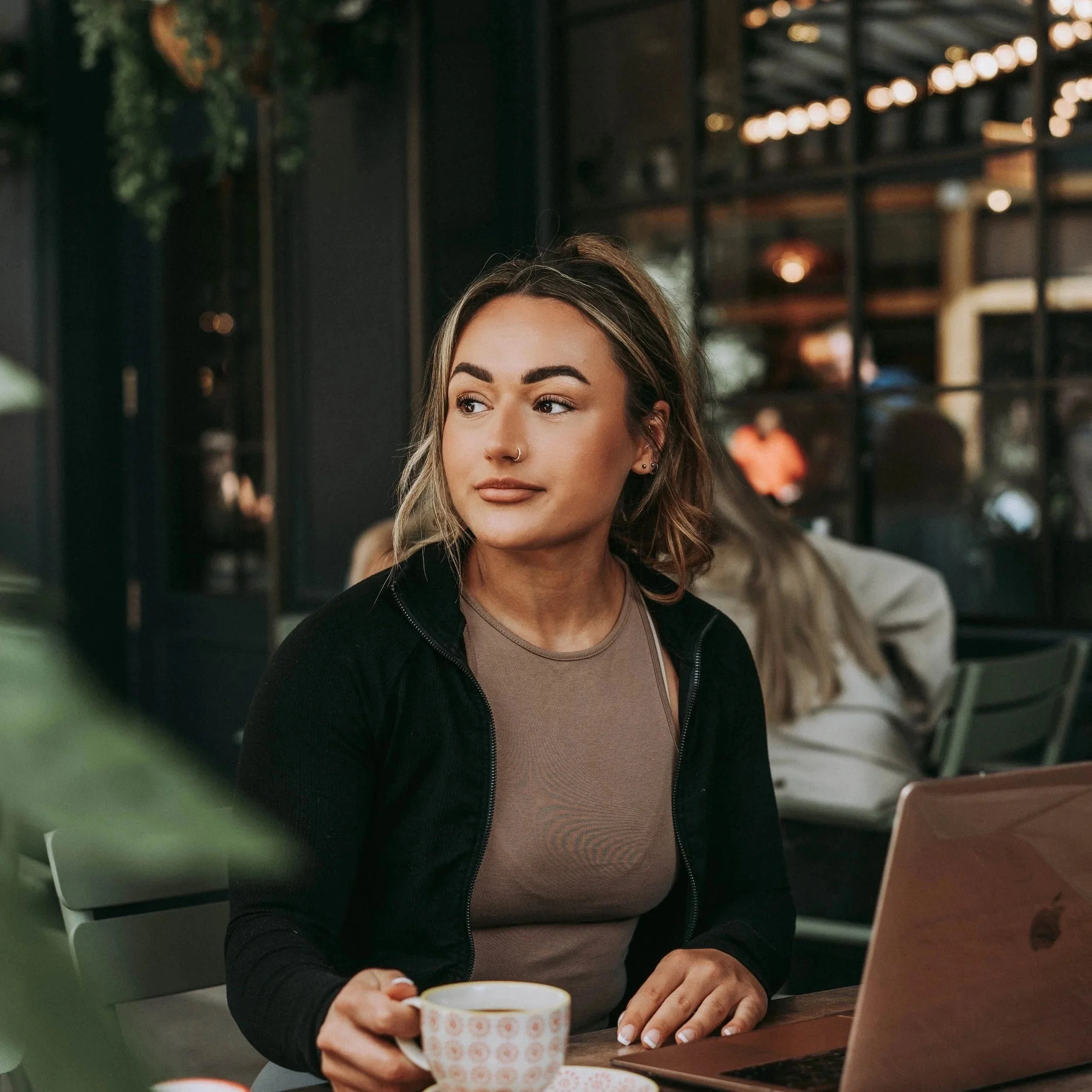 A woman sitting at a table in a cafe or coffee shop, holding a coffee cup, with a laptop open in front of her, looking to the side.