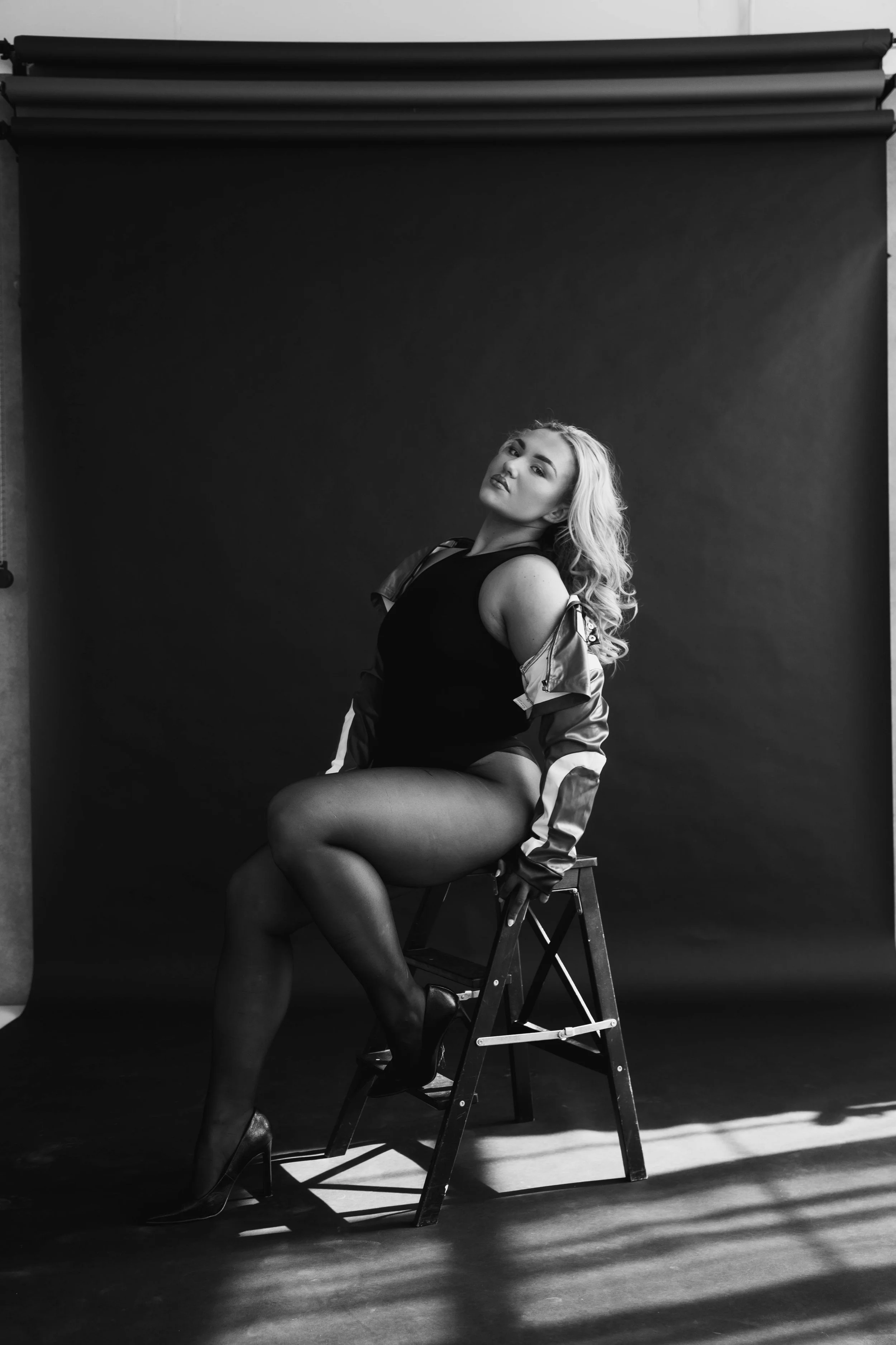 A woman with long wavy blond hair sitting on a high stool, posed with one leg crossed over the other, in front of a black backdrop. She is wearing a black top, shiny jacket, stockings, and high heels, with a confident expression.